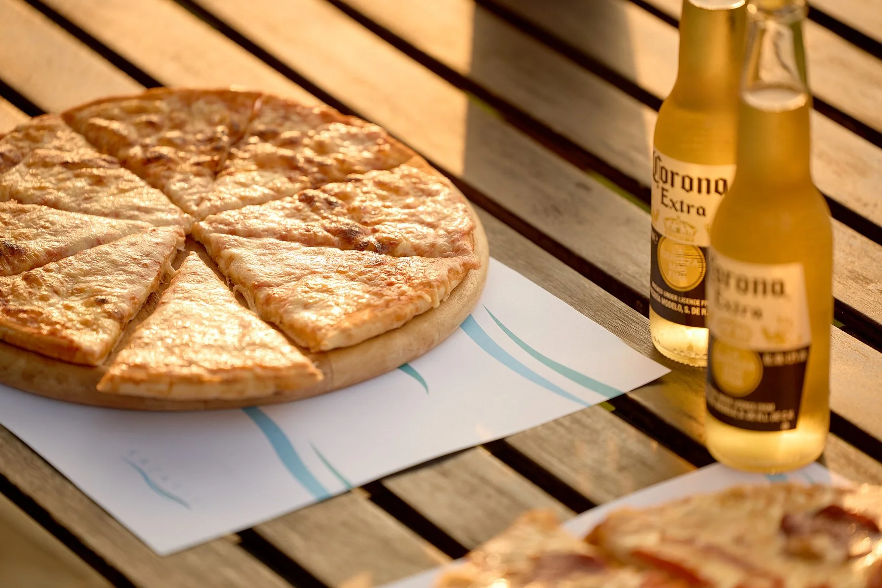 A pizza with cheese on a wooden table with two bottles of Corona beer nearby.