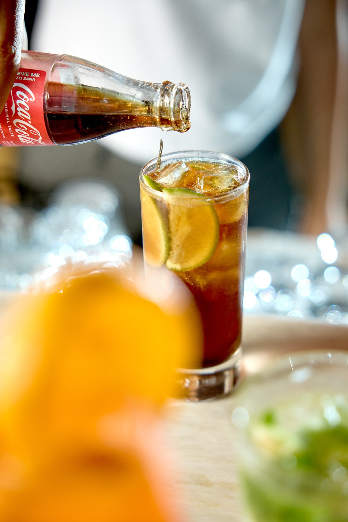 A glass of iced Coca-Cola with lime slices, with a Coca-Cola bottle pouring into it, on a blurred background.