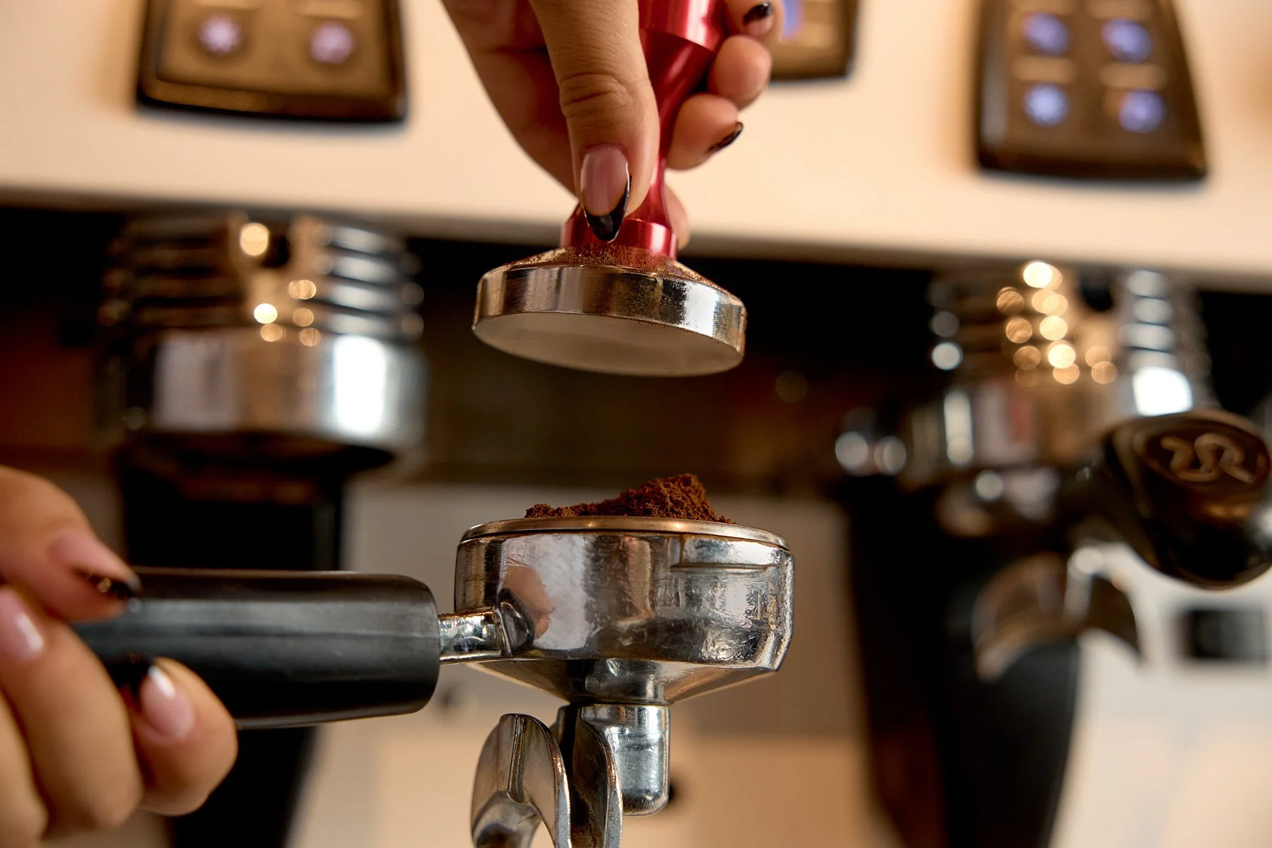Close-up of a barista's hand pressing a tamper into fresh ground coffee in a portafilter, with espresso machine controls in the background.