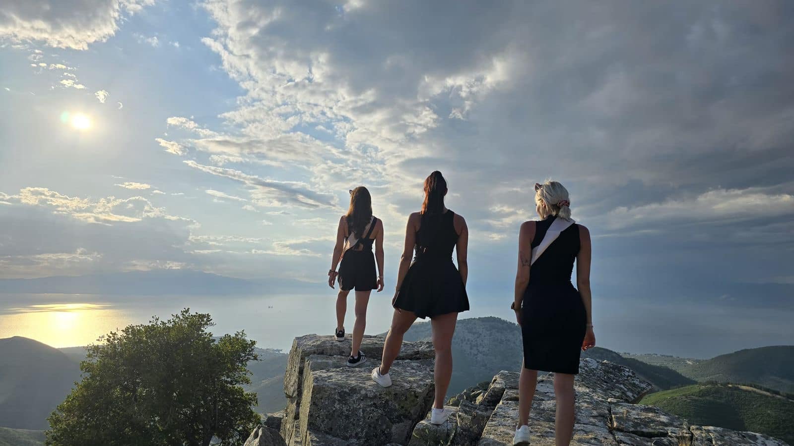 Three women standing on rocks on a mountain overlooking a body of water during sunset.