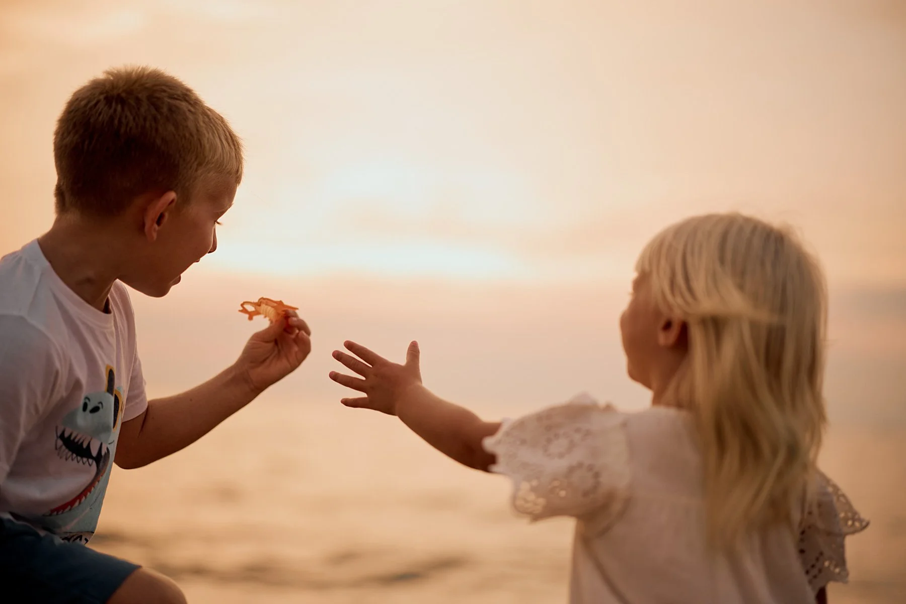 A boy and girl playing together near the water during sunset, with the boy giving the girl a small toy.