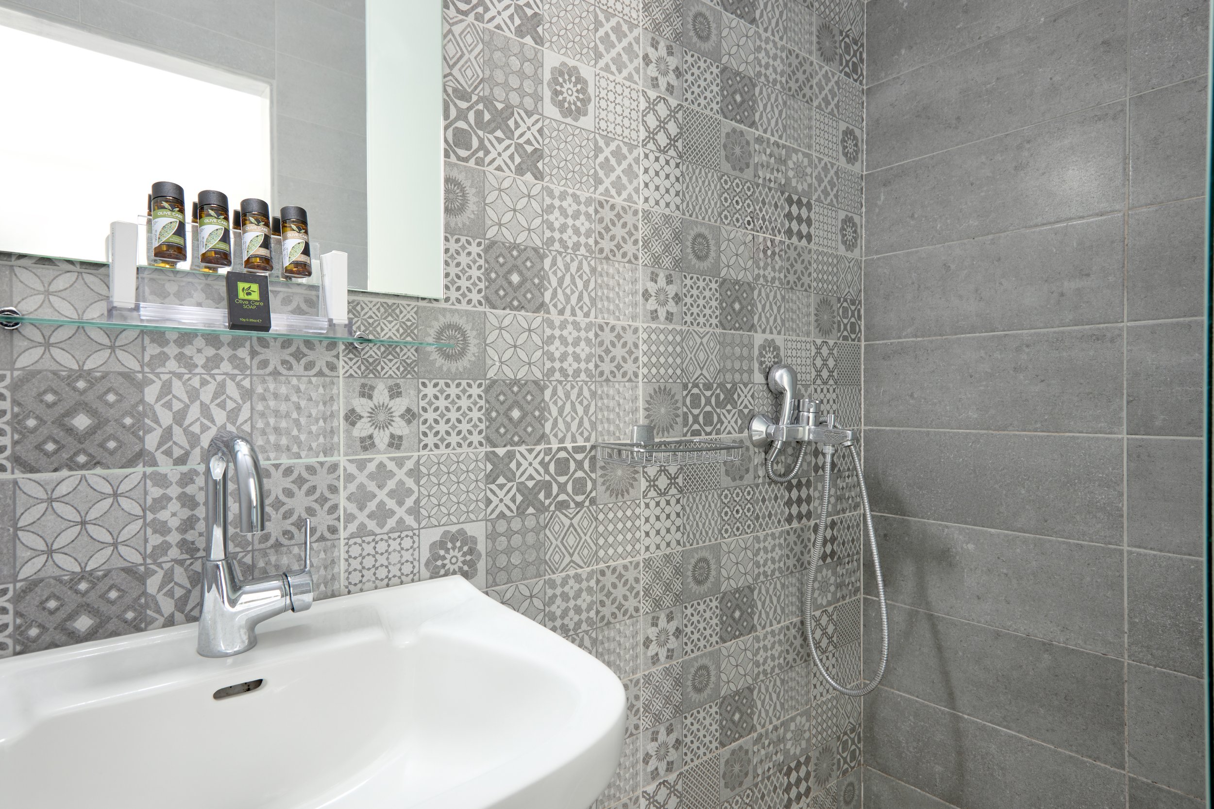 A modern bathroom with a small white sink, patterned gray tiles on the wall, a mirror above the sink, and a shower area with gray tiles and a handheld showerhead.