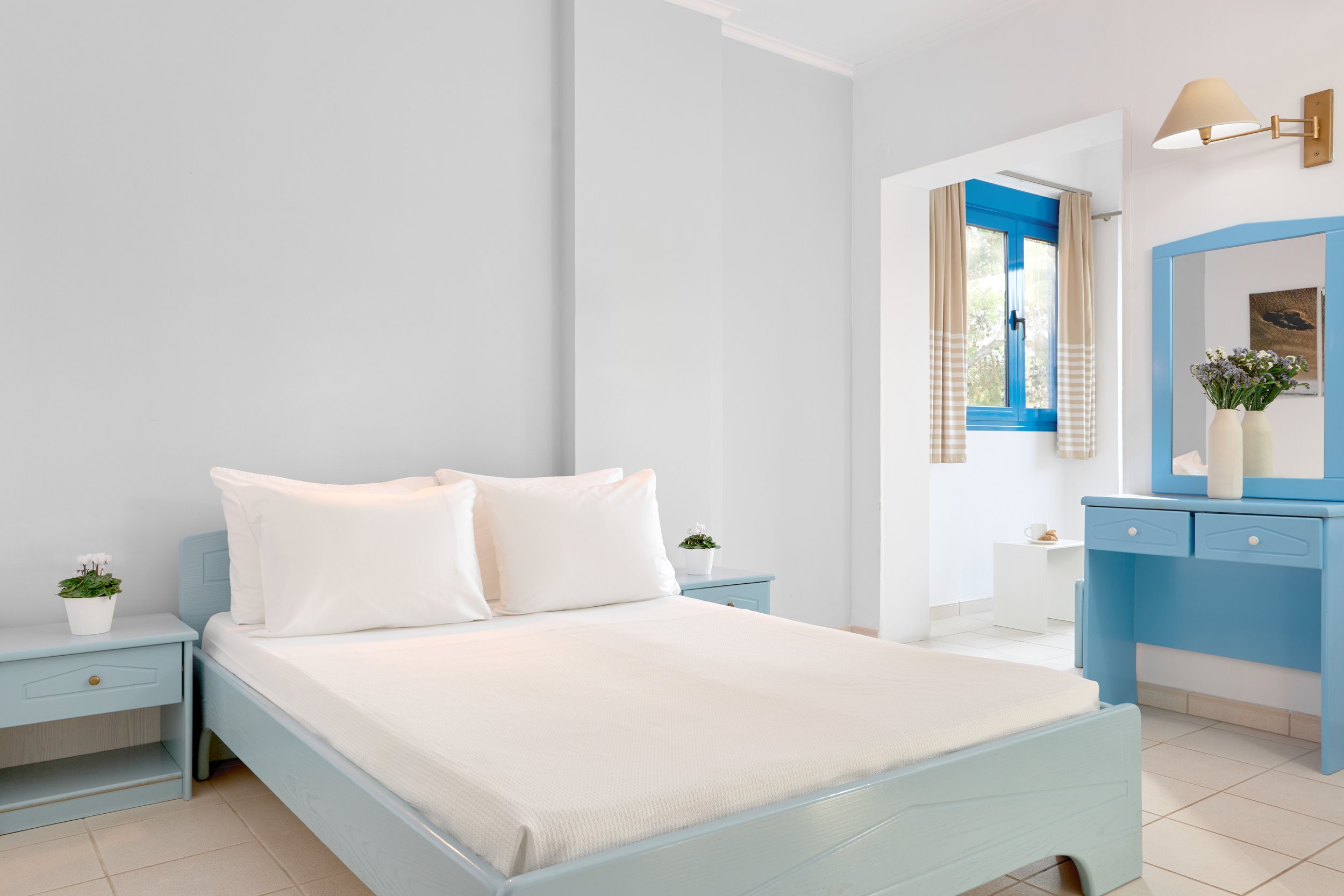 A bright bedroom with white walls and bedding, two white pillows, and blue furniture including two nightstands and a dresser with a mirror. The room has a window with beige checkered curtains and blue trim, decorated with vases of flowers.