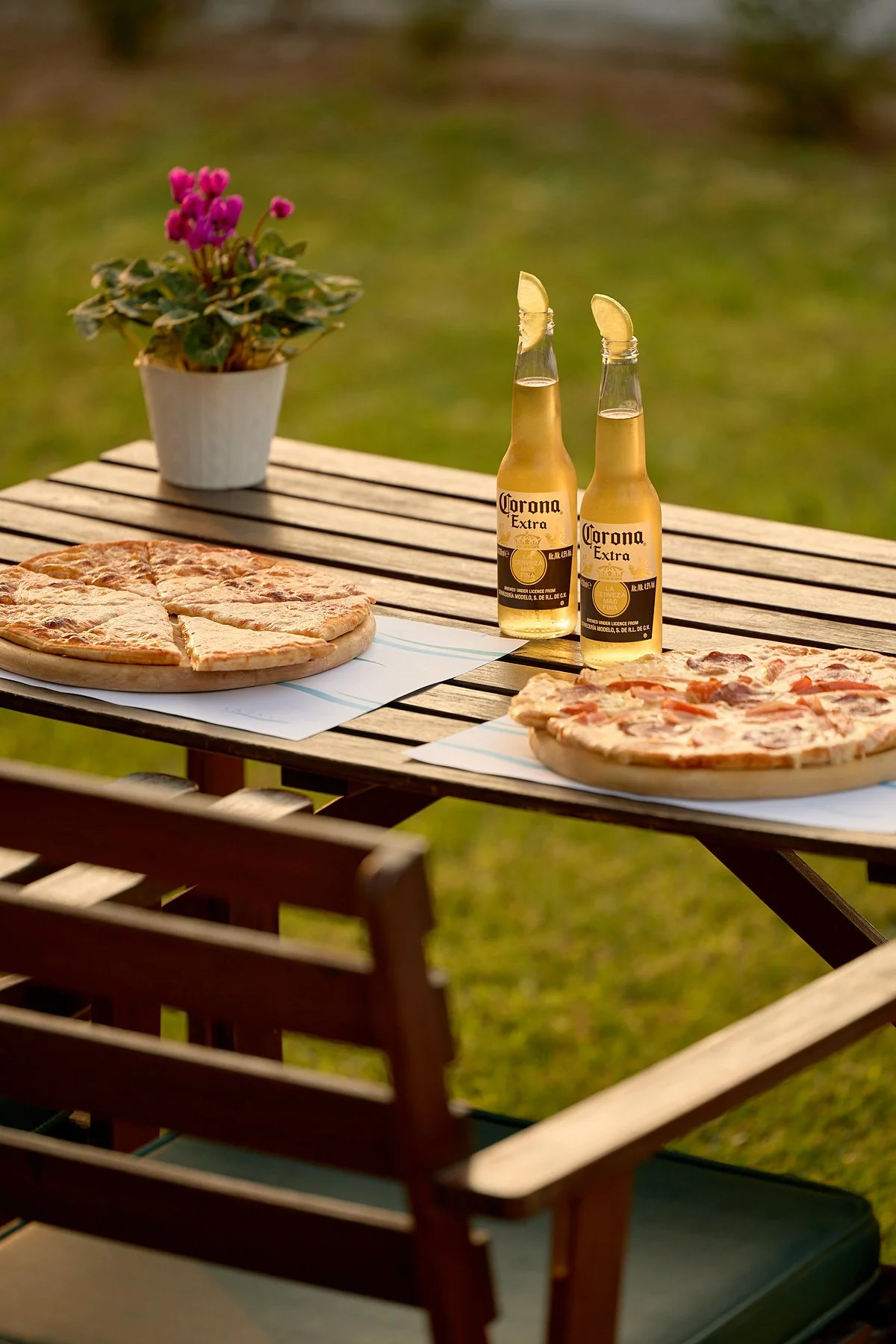 Two bottles of Corona beer with lime wedges on a patio table next to two pizzas, one with cheese and the other with toppings, and a potted purple flower in a white pot in the background, outdoors on a sunny day.