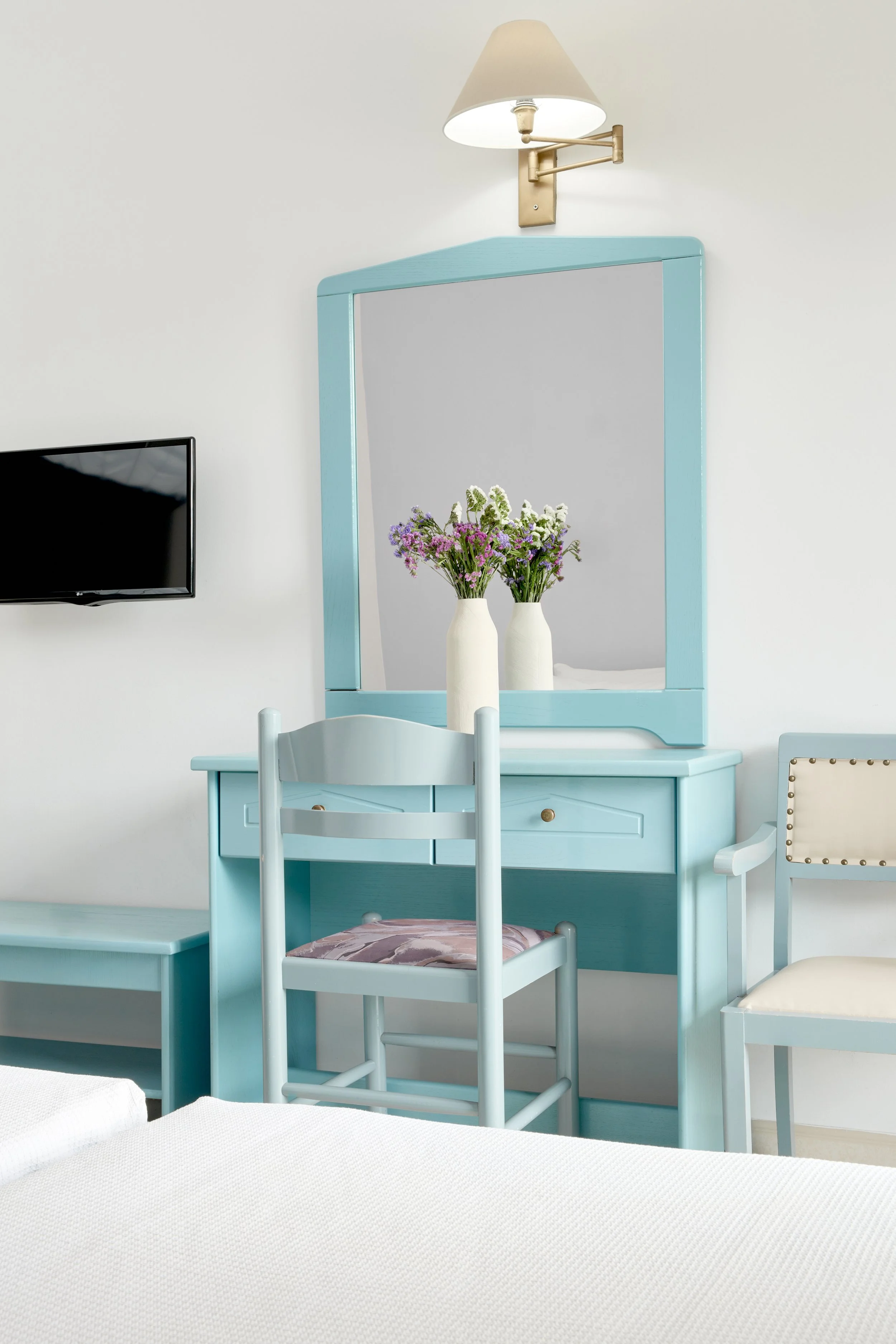 A bedroom with a blue vanity table, a matching chair with a patterned cushion, and a large mirror. Two white vases with flowers are on the vanity. A wall-mounted TV and a wall sconce light fixture are also visible.