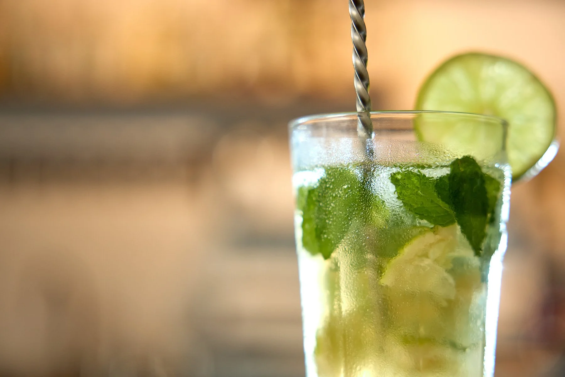 Close-up of a tall glass of mojito with fresh mint leaves, lime slices, ice, and a metal straw, with a lime wheel garnish.