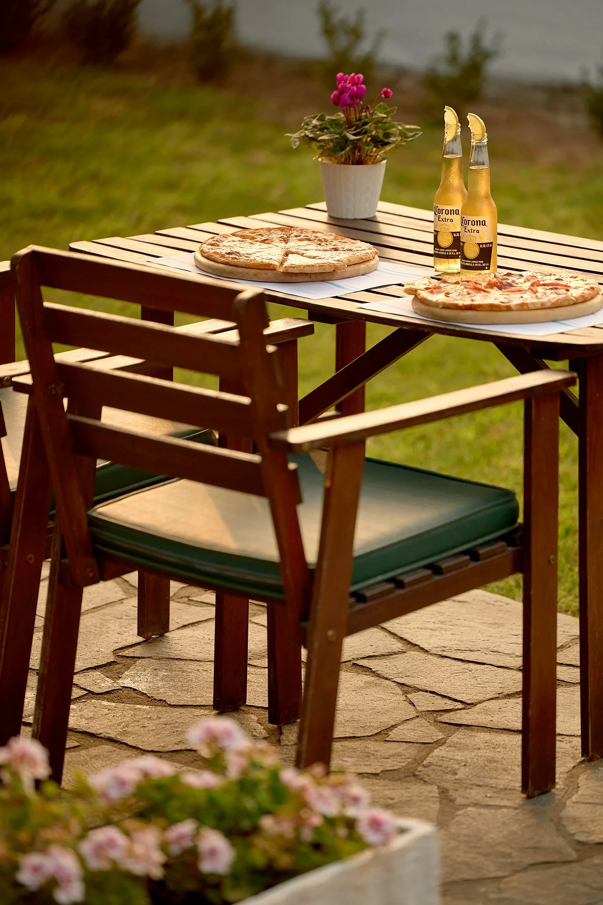 An outdoor wooden table with two pizzas, two bottles of Corona beer with lemon wedges, a potted plant with pink flowers, and three chairs on a stone patio, overlooking a grassy area and a body of water.