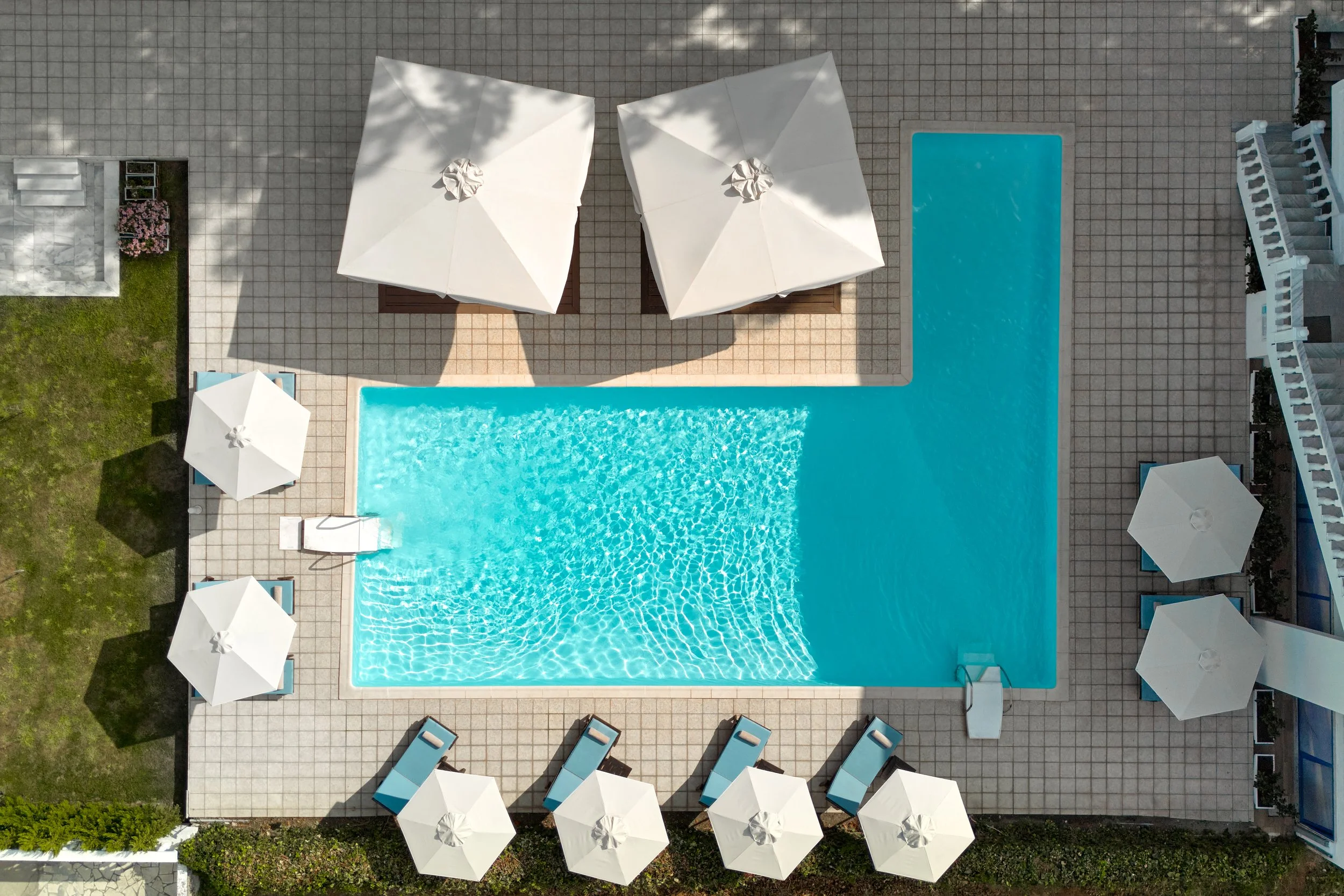 Aerial view of a swimming pool area with blue water, surrounded by lounge chairs and white umbrellas on a tiled deck, with a grassy area and a building with a staircase nearby.