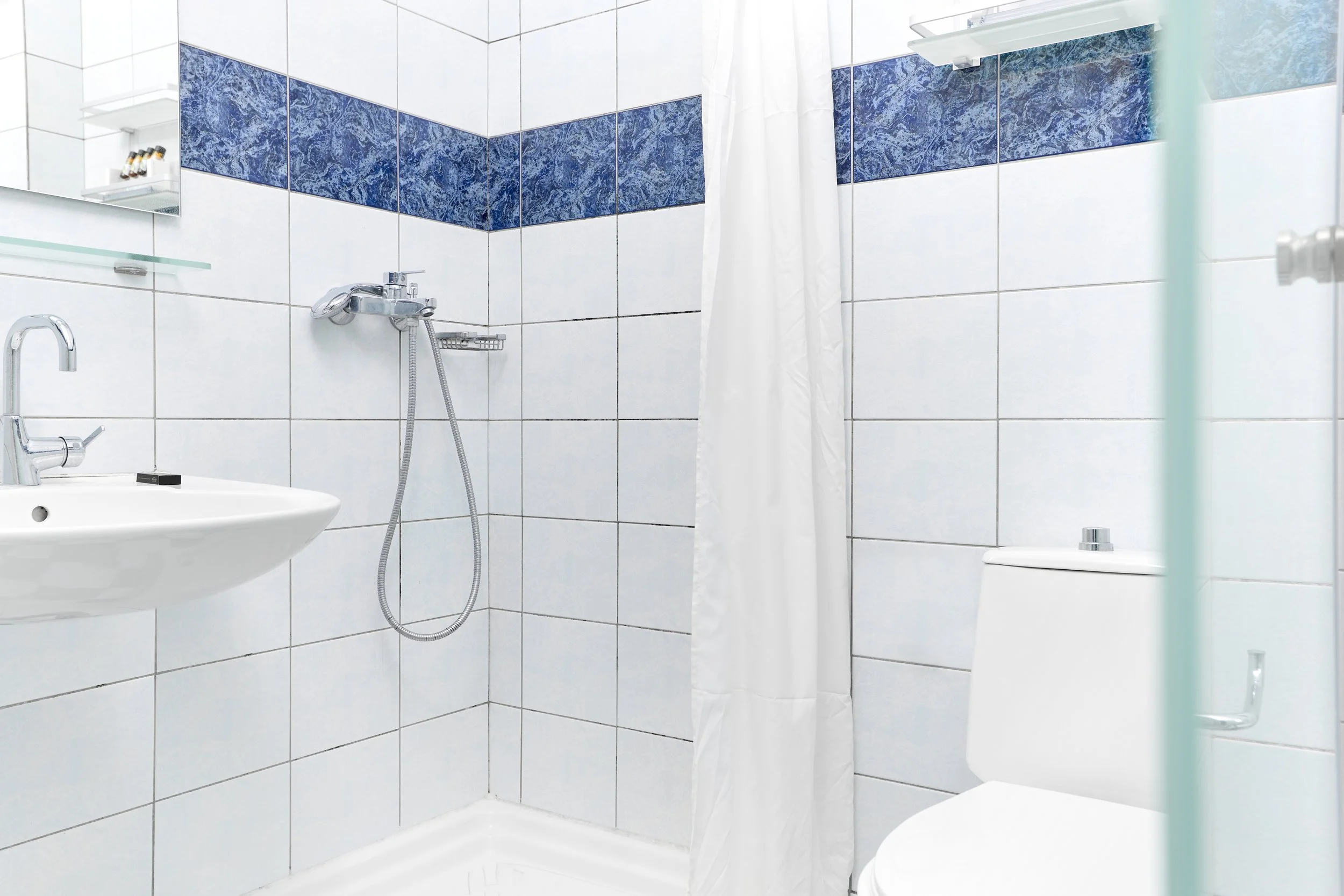 A small white-tiled bathroom with a shower area, a white toilet, a wall-mounted mirror, a small glass shelf with toiletries, and a shower curtain.