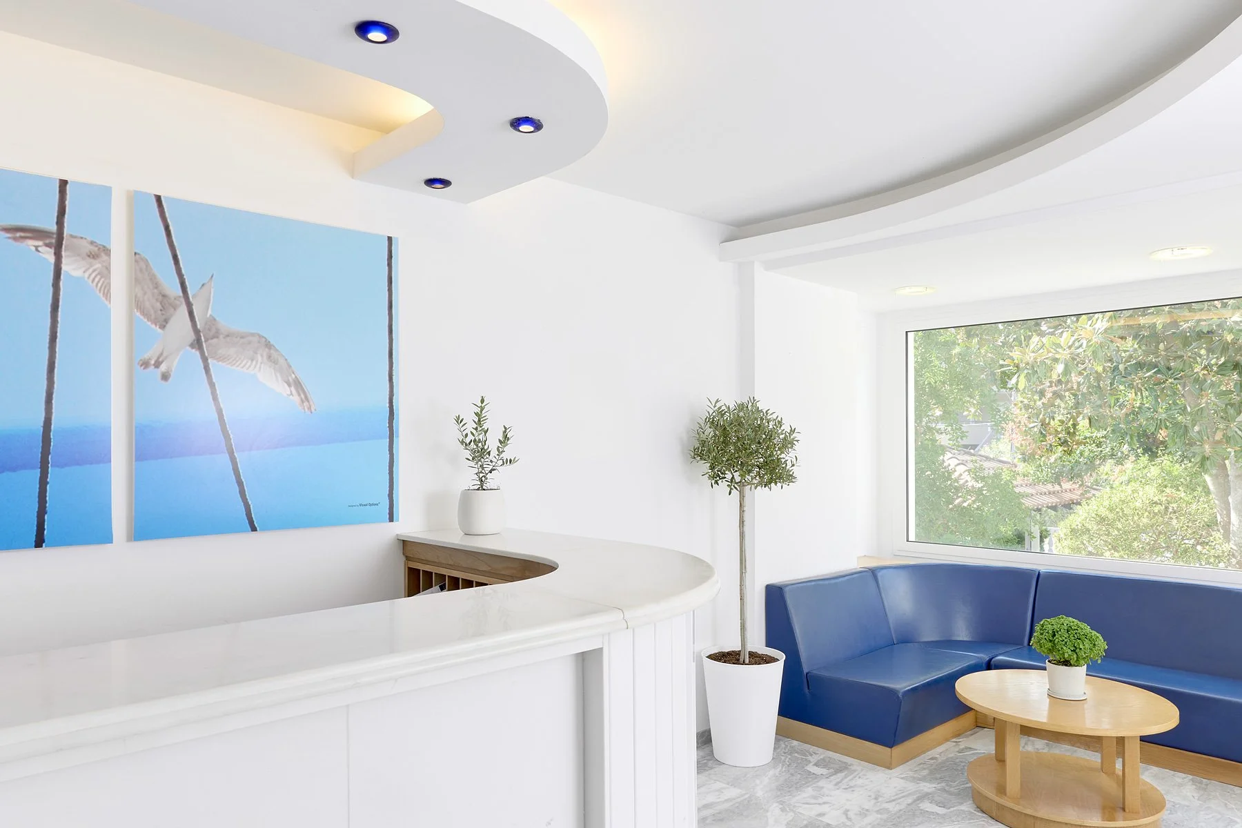 Modern interior lobby with white walls, blue seating, potted plants, large window with trees outside, and a abstract bird artwork on the wall.