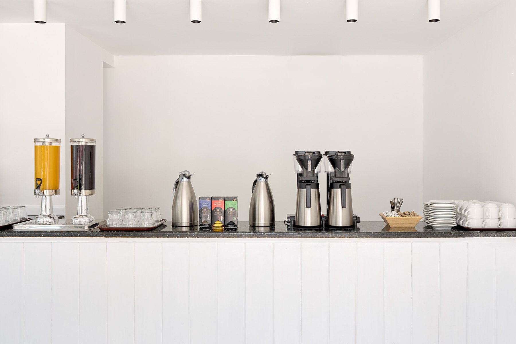 Coffee and juice station with dispensers, pots, cups, and stirrers on a black countertop against a white wall.