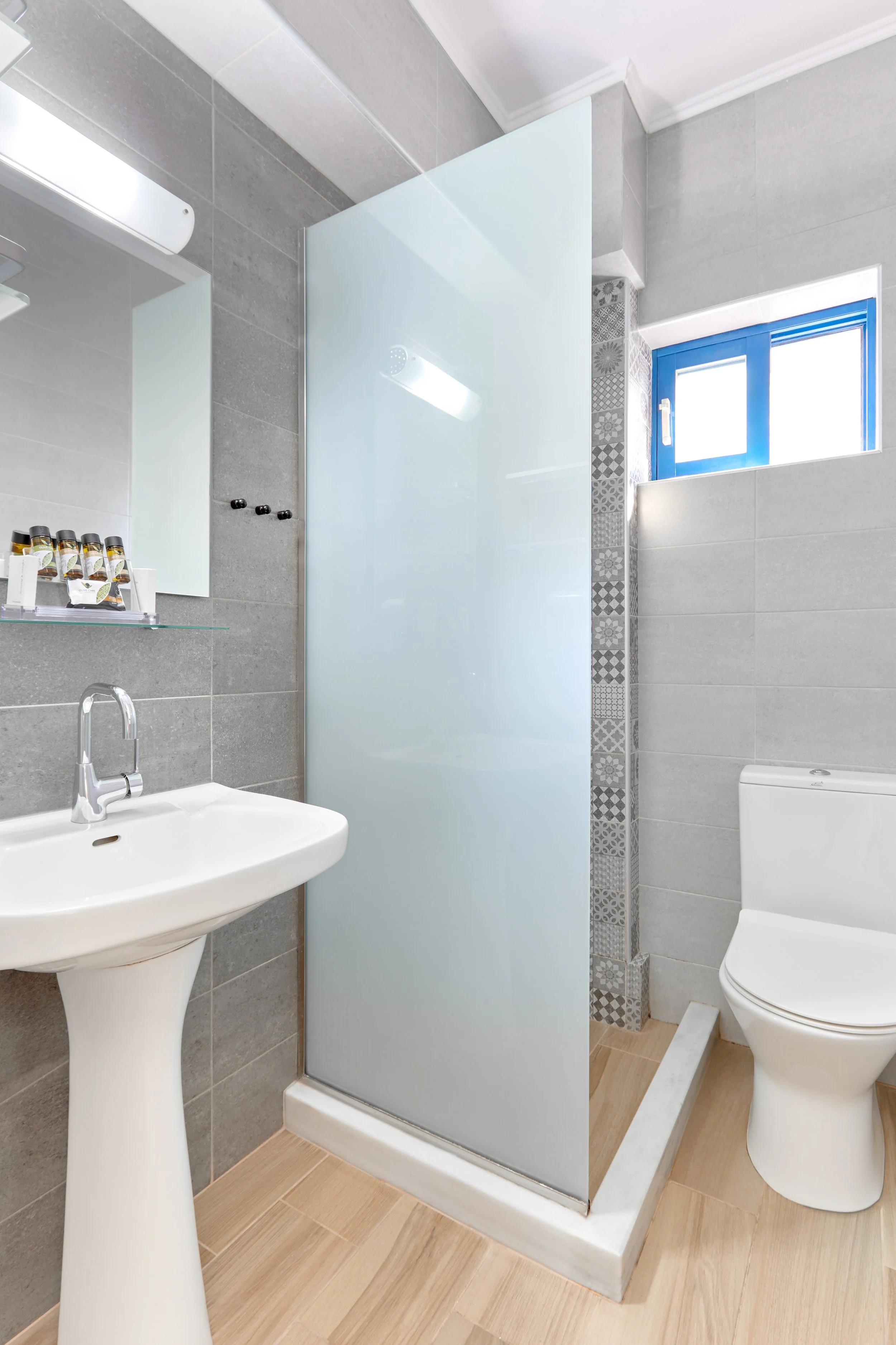 A small modern bathroom with gray tiled walls, a white pedestal sink, a mirror, a glass shelf with toiletries, a white toilet, a frosted glass shower partition, and a small window with blue trim.