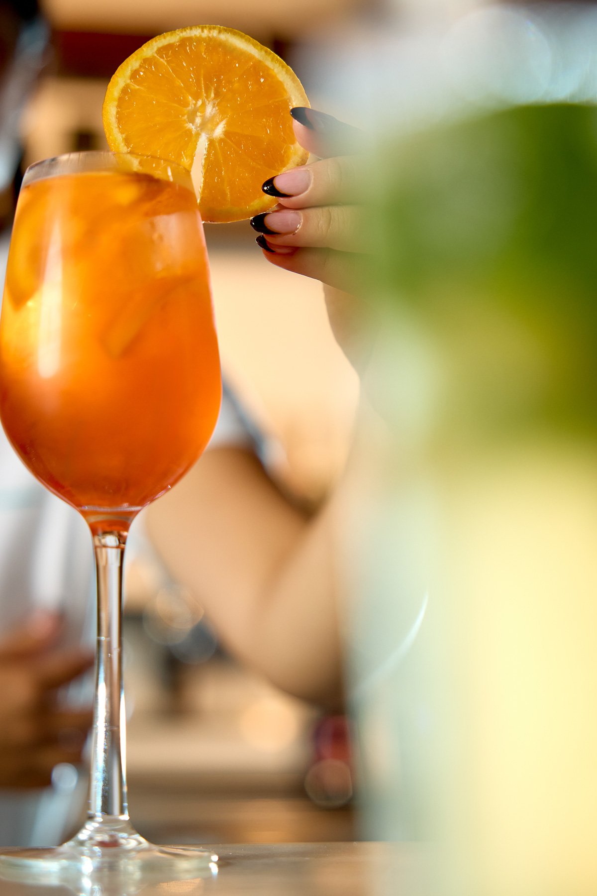 A cocktail with orange slices in a tall glass, garnished with a slice of orange, on a counter with a blurred background.