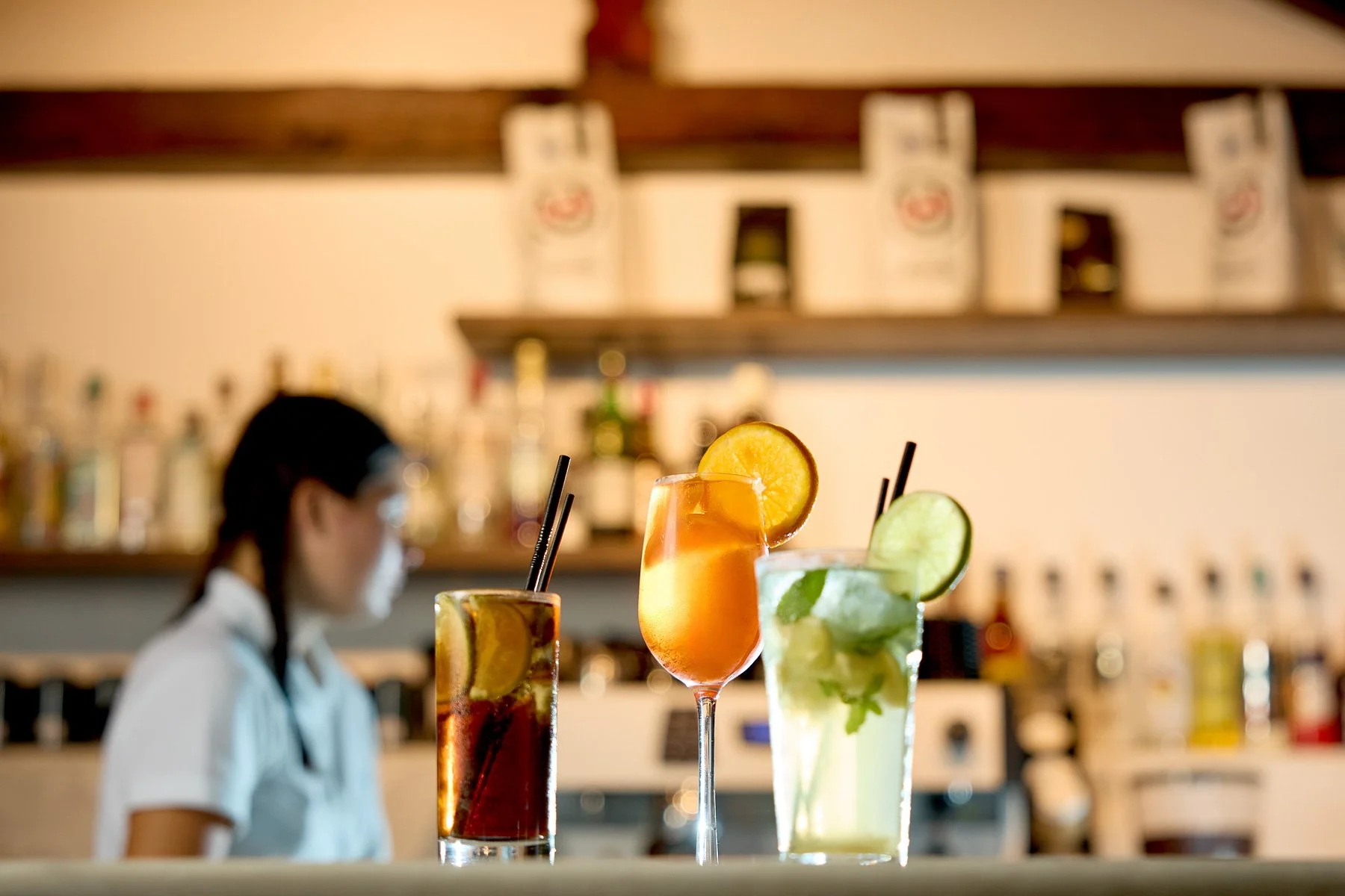 Three colorful cocktails on a bar counter with blurred background of a bartender and shelves of bottles.