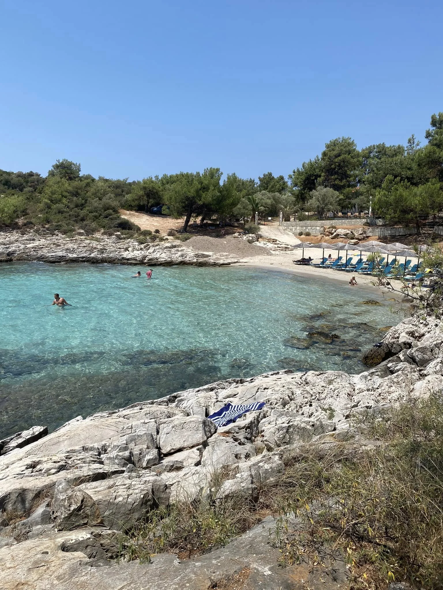 A scenic beach with clear turquoise water, rocky shoreline, and green trees in the background. Several lounge chairs with umbrellas are on the sandy beach, and a few people are swimming and relaxing.