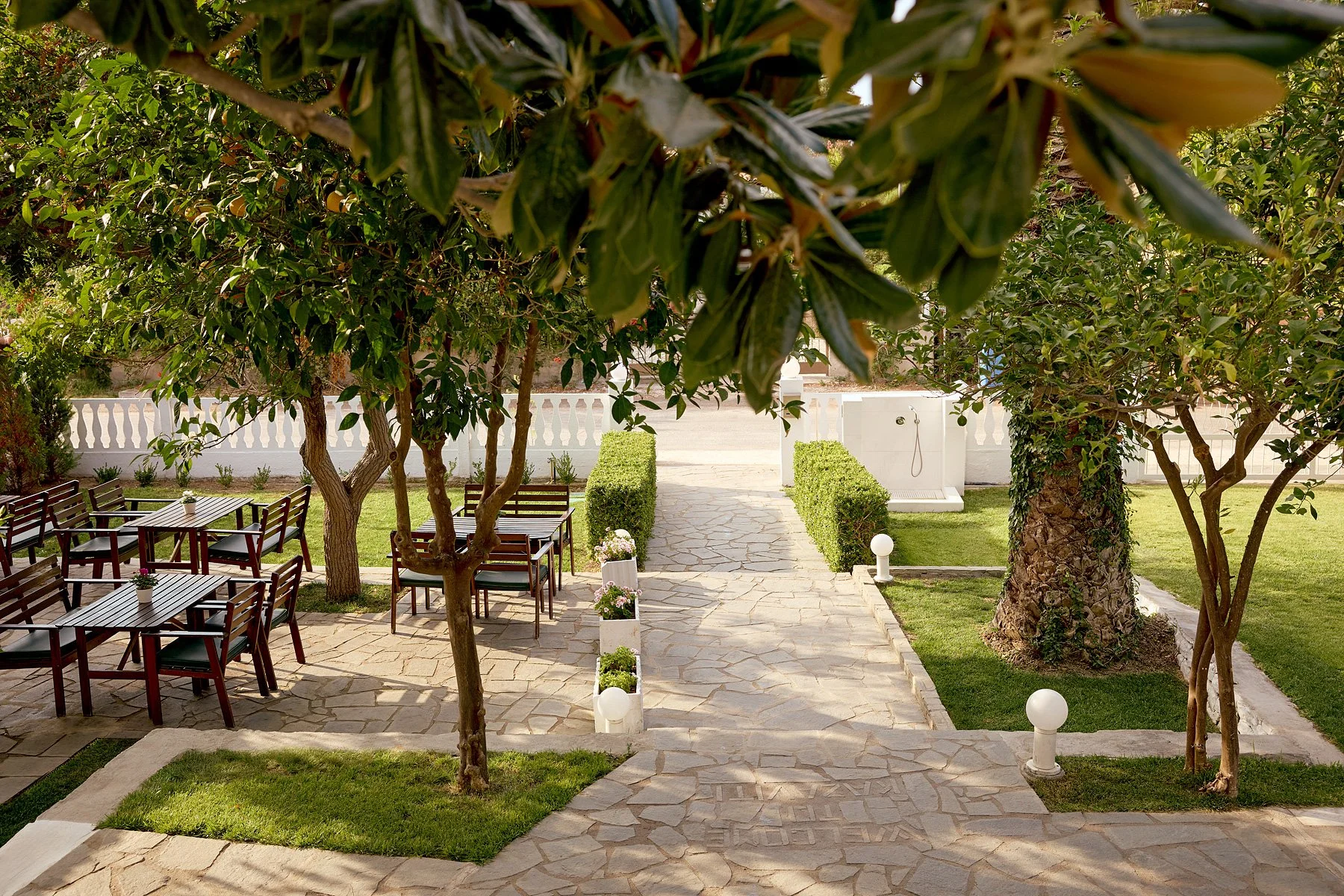 Outdoor garden with stone pathway, trees, and seating area with wooden tables and chairs under the shade.