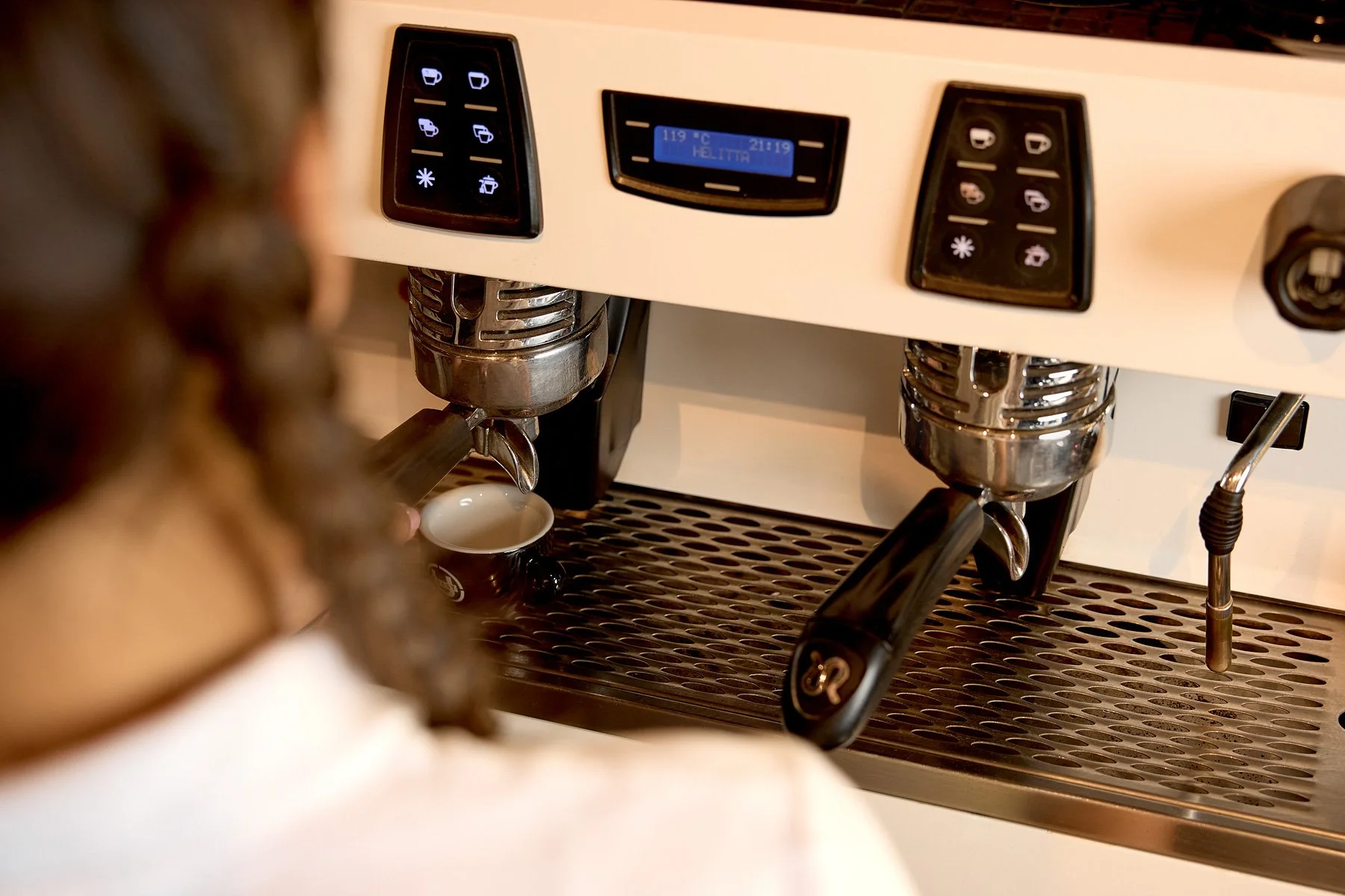 A barista making coffee with a dual espresso machine, filling a small cup with espresso, with digital displays and control panels.