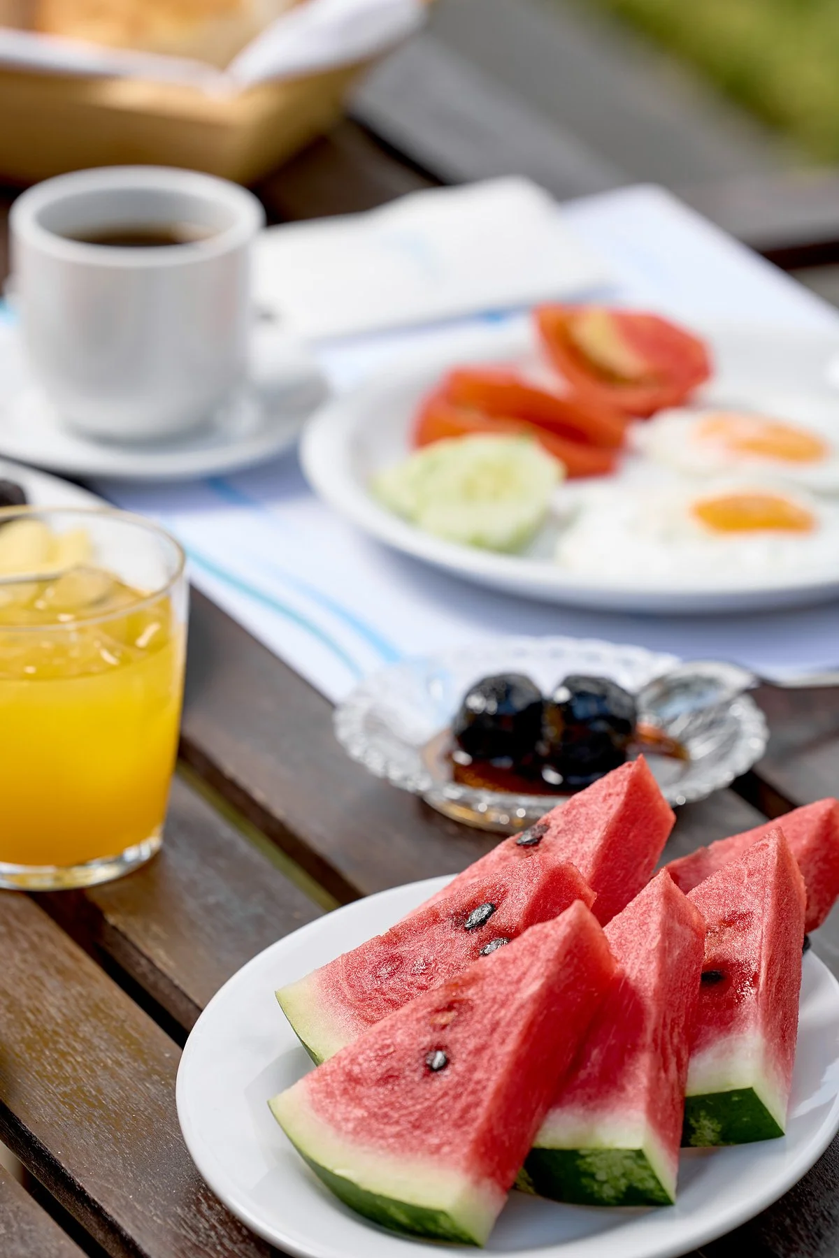 A breakfast spread on a wooden table includes slices of watermelon on a white plate, a glass of orange juice, a cup of coffee, and a plate with eggs, tomatoes, and cucumber slices.