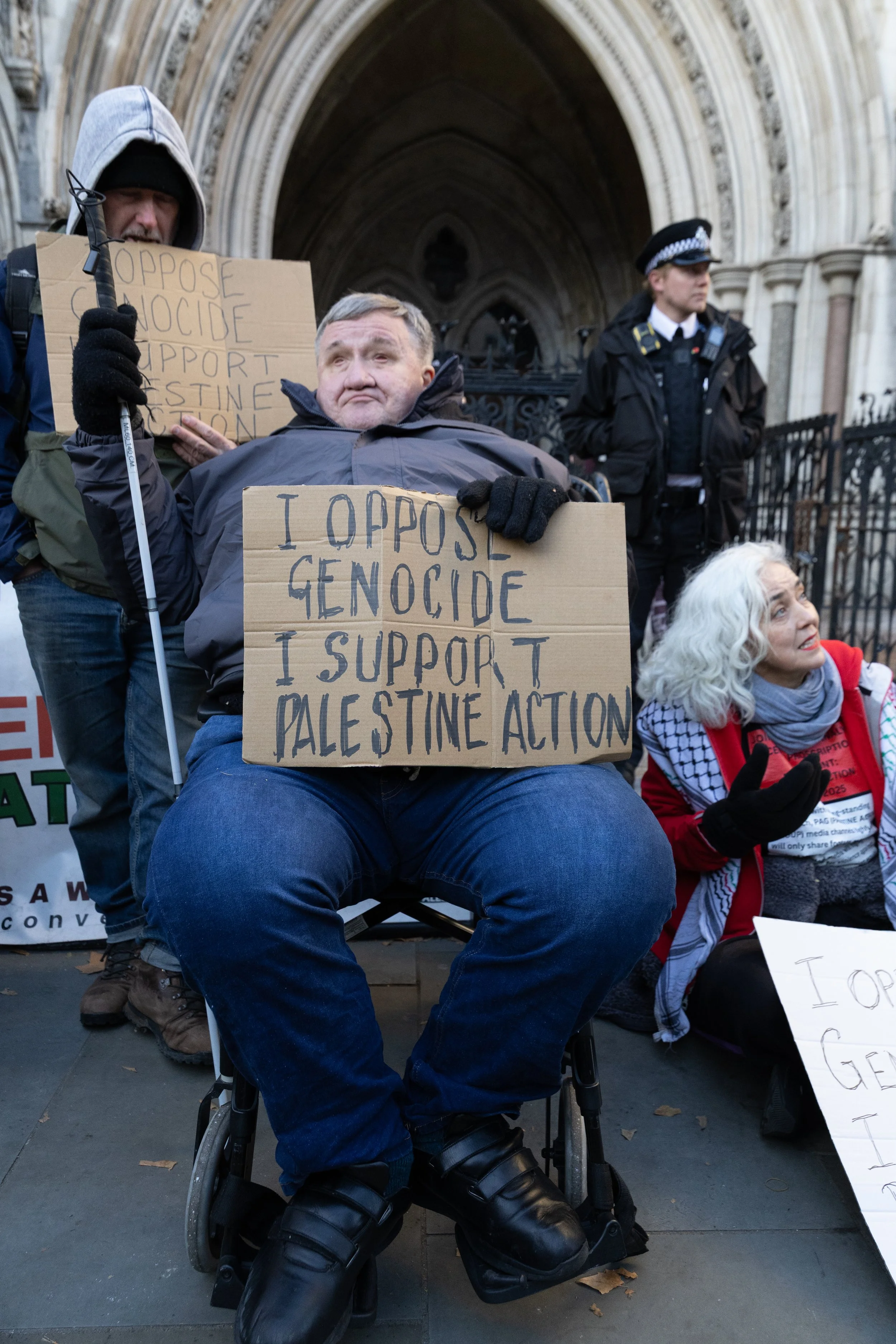 A man in a wheelchair holding a cardboard sign that says 'I oppose genocide I support Palestine action,' part of a protest outside a historic building with people and police in the background.