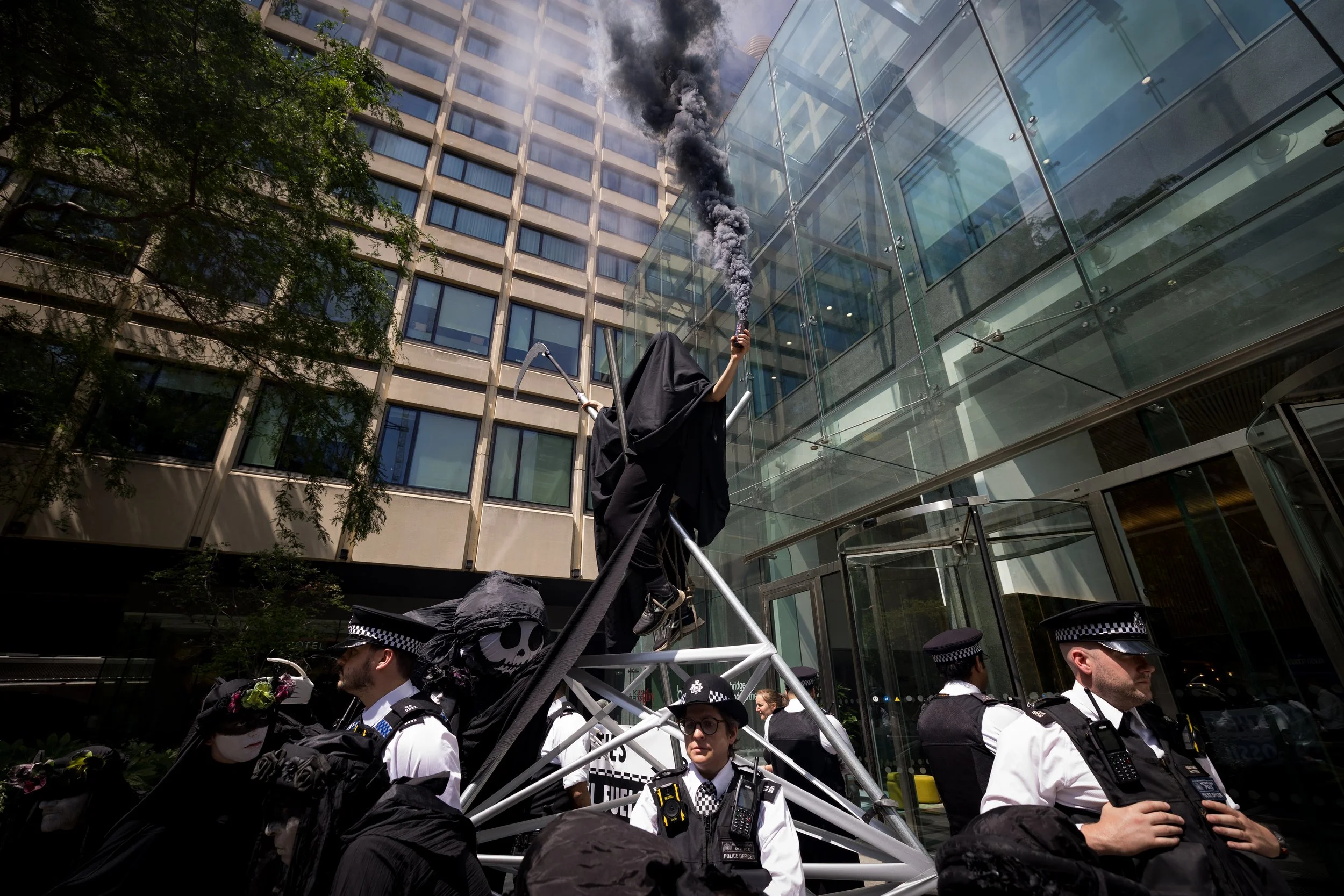 A protest scene with a large puppet of the Grim Reaper holding a smoke bomb, surrounded by police officers, in front of a modern glass building.