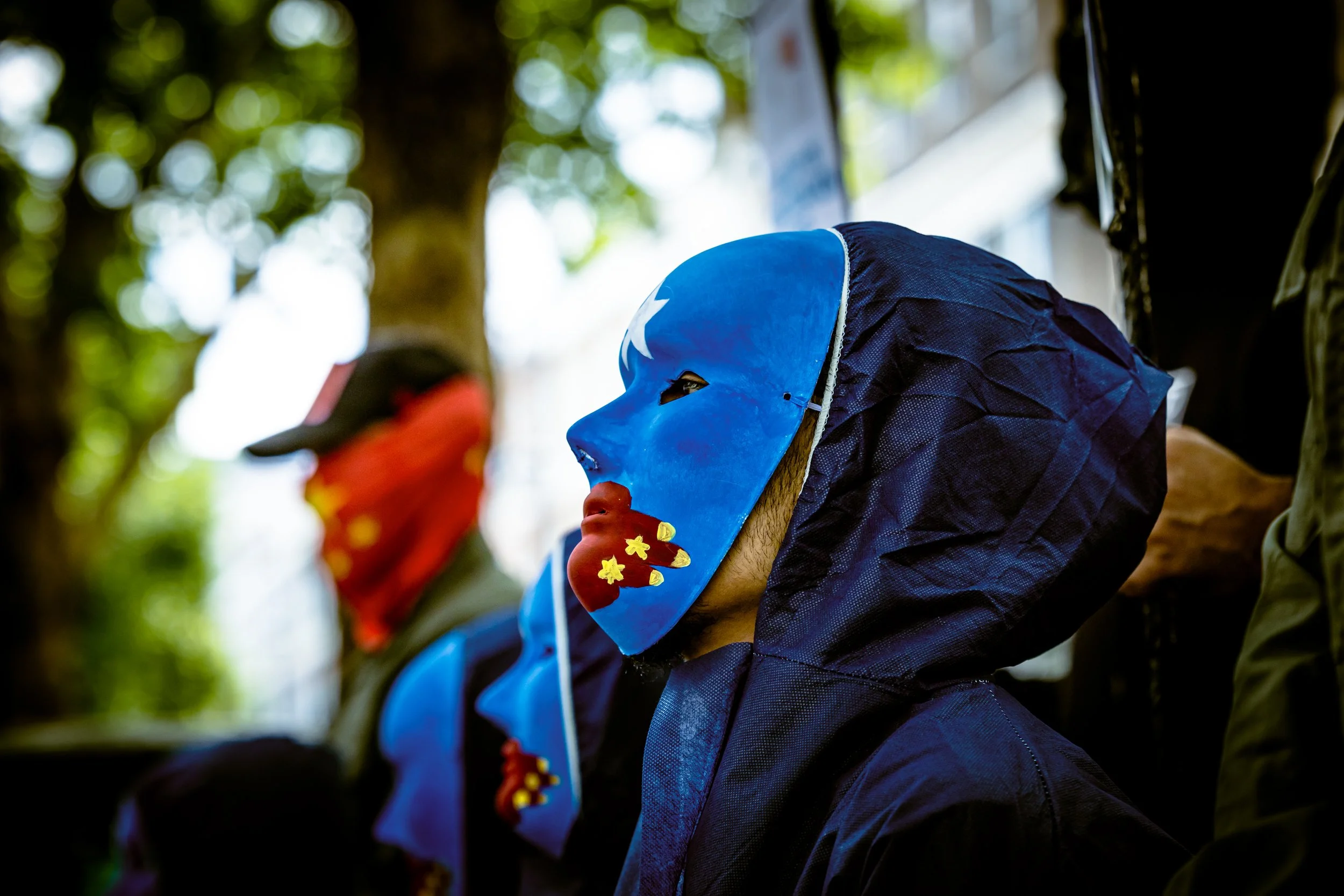 A person wearing a blue mask with white and red decorative elements, and a dark blue jacket, appears to be participating in a celebration or event outdoors, with blurred green foliage in the background.