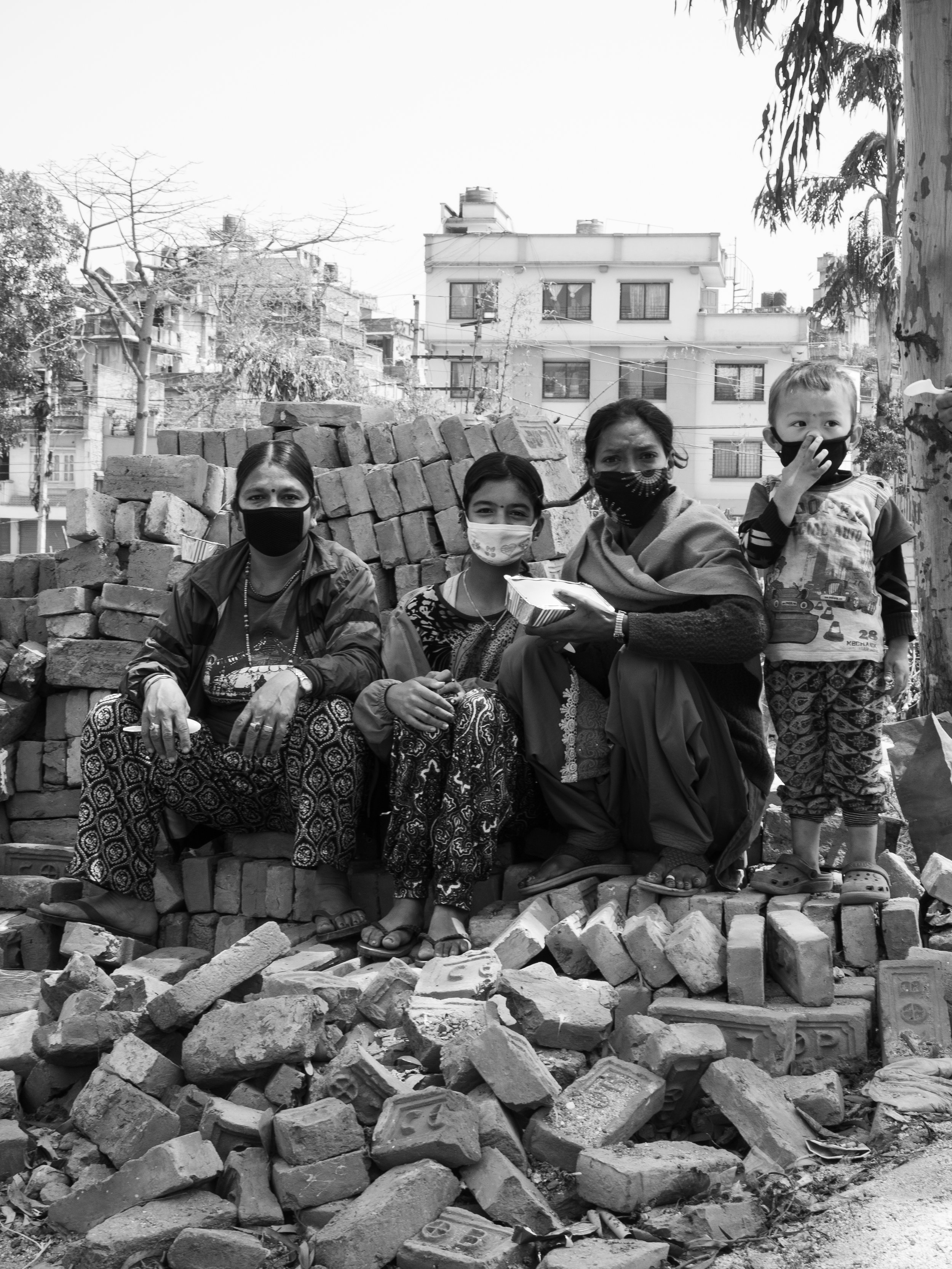 Four women and one young boy wearing face masks, sitting and standing on a pile of bricks with buildings in the background.