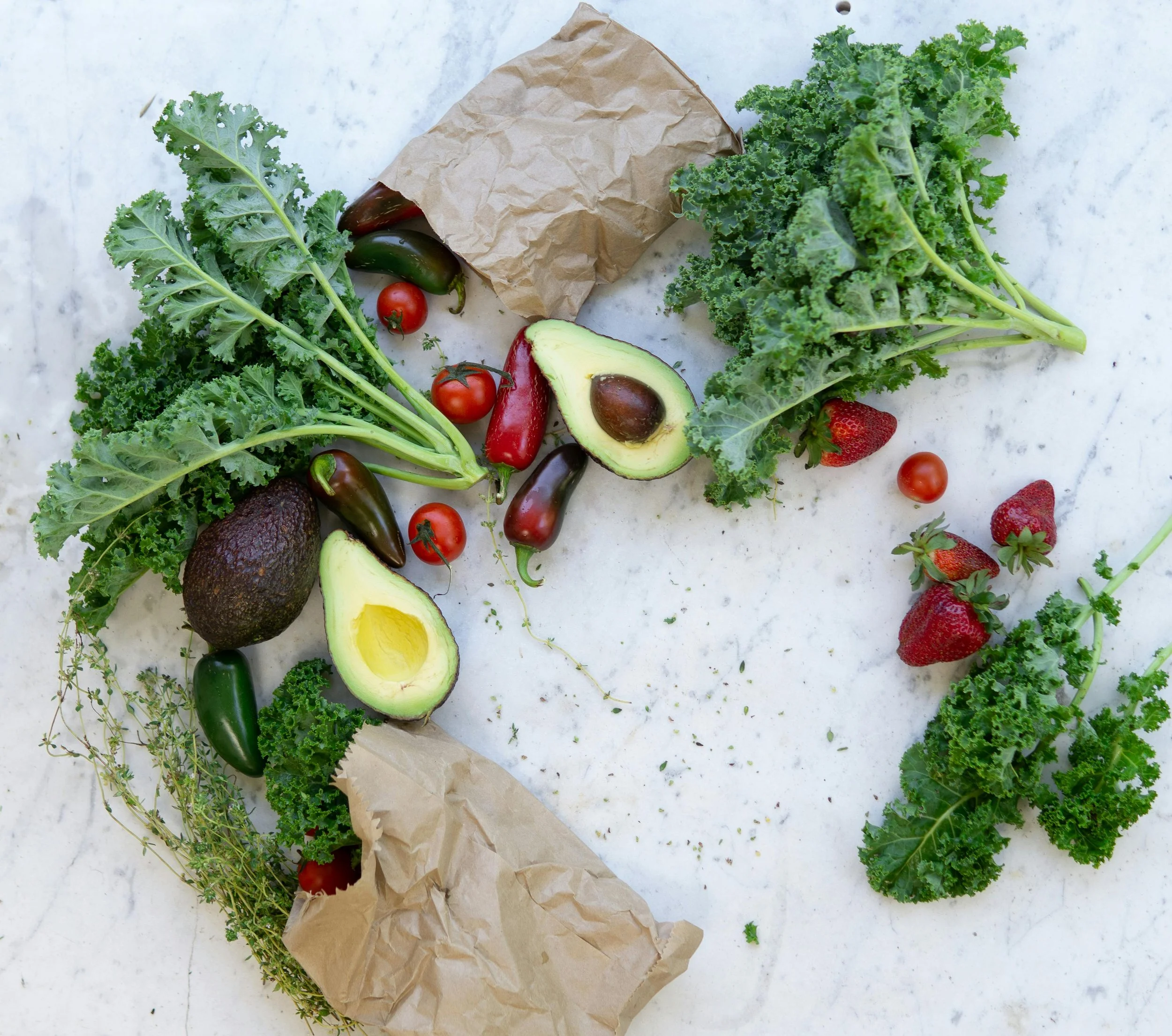 Fresh green kale, avocado halves, cherry tomatoes, strawberries, and herbs on a white surface.