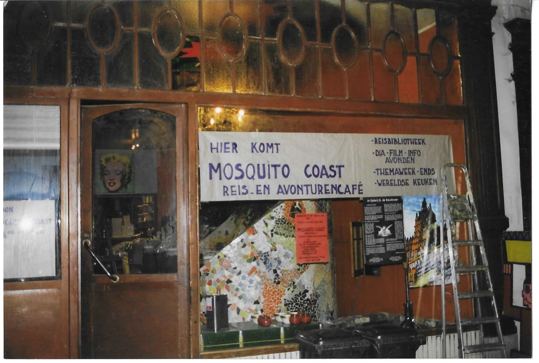 Uitzicht op de ingang van een reis- en avonturen café genaamd Mosquito Coast met een handgeschreven banner en een ladder voor de deur.