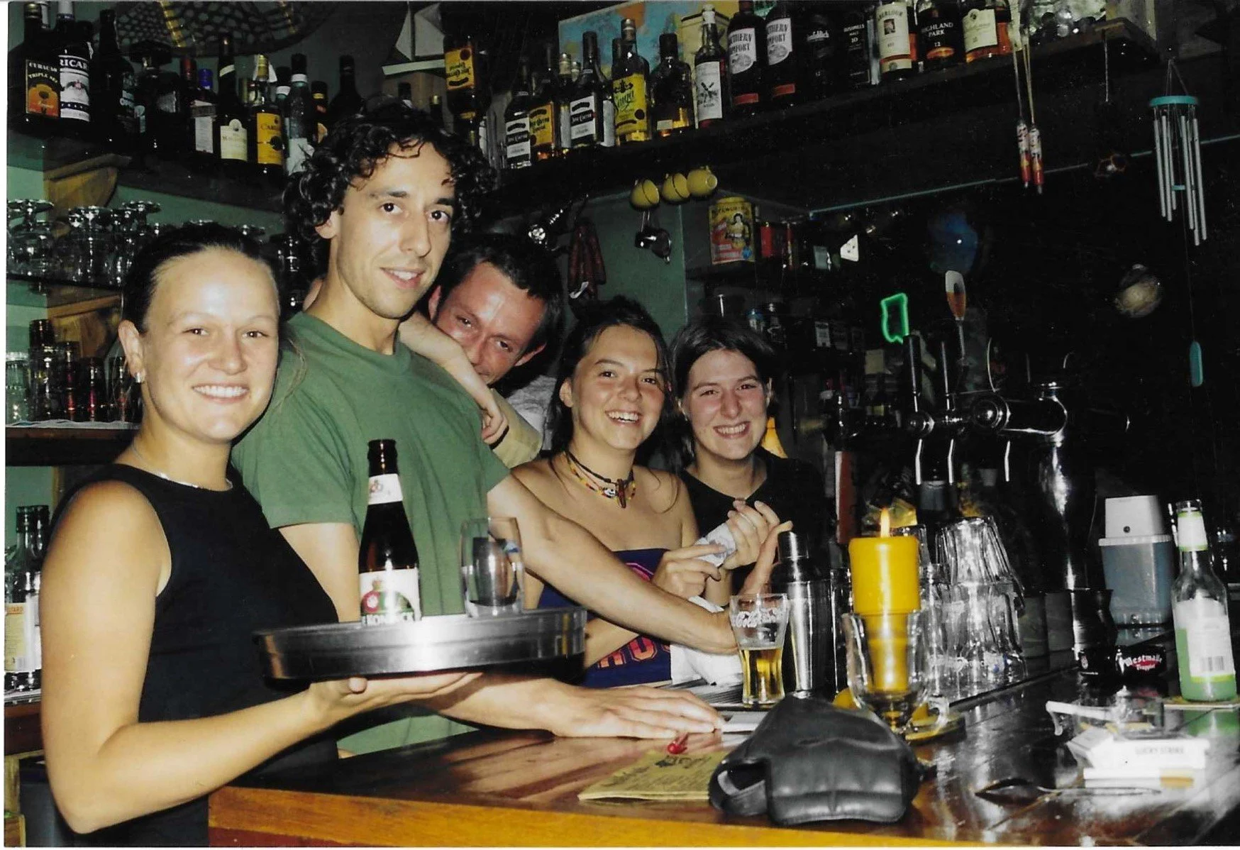 Vijf mensen die in de nineties in bar & restaurant Mosquito Coast in Gent staan, met drankjes en glazen op de tafel, en een achtergrond vol flessen en schappen.