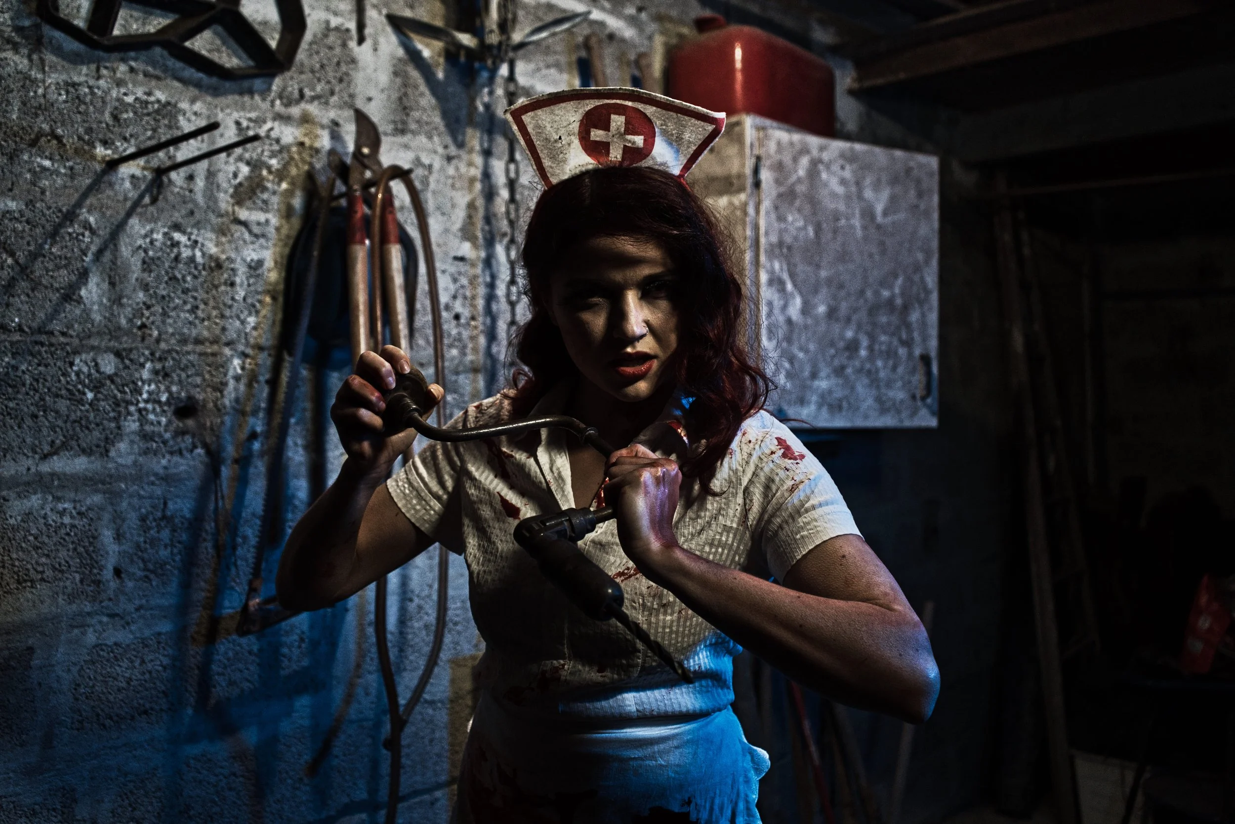 Une femme portant un uniforme d'infirmière avec un symbole de croix rouge, tenant un outil dans un environnement sombre et sale, avec des outils suspendus au mur en arrière-plan.