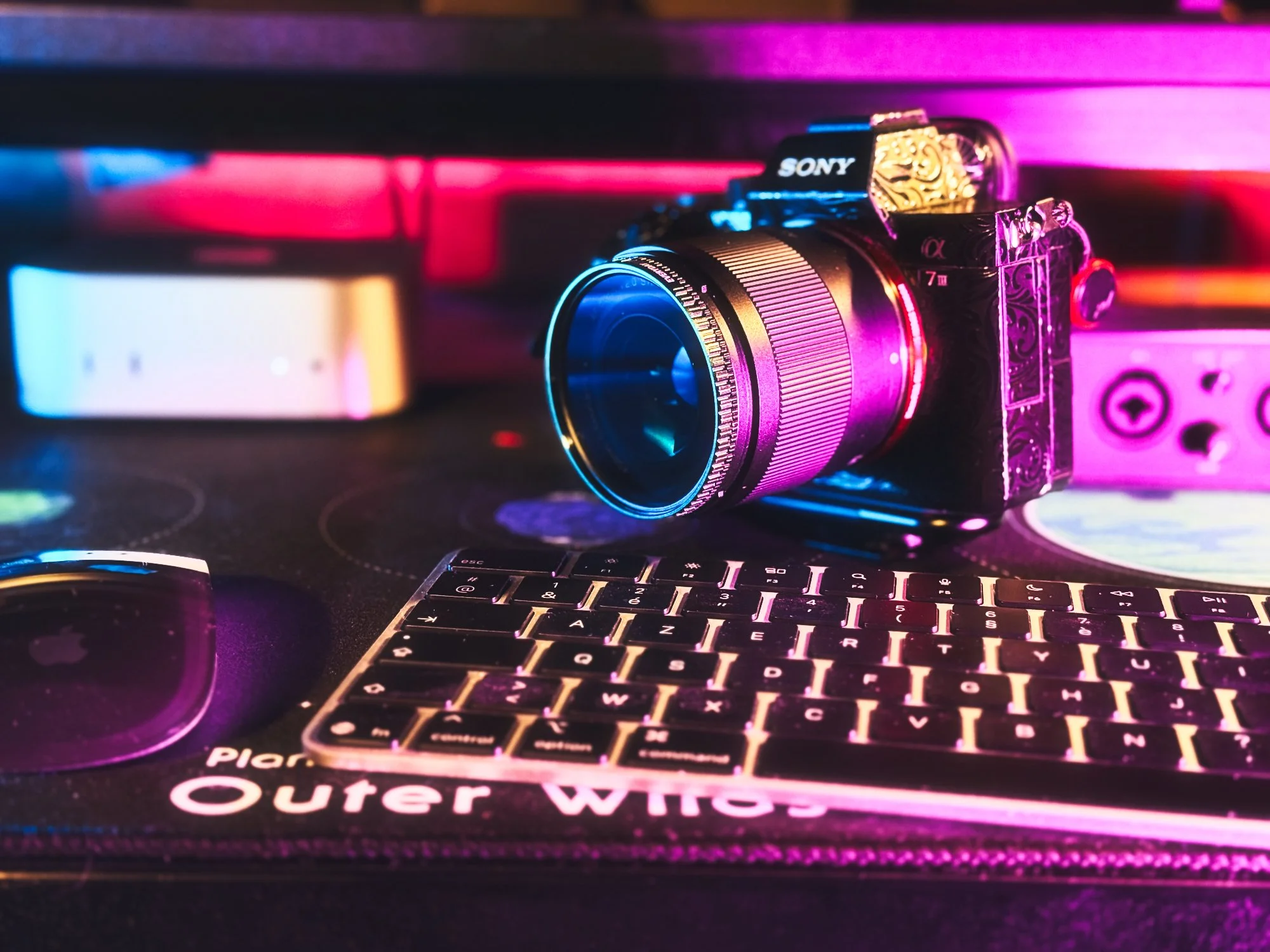 Appareil photo Sony, clavier et souris Apple sur un tapis de bureau. Éclairage coloré en bleu et rose.