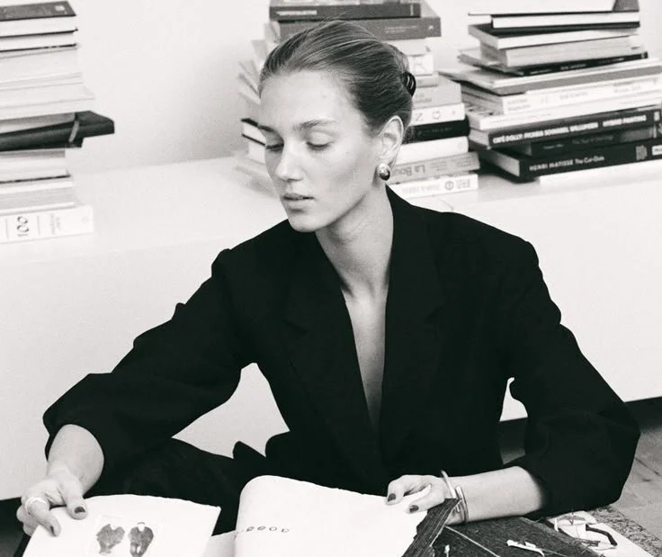Schöne Frau mit hochgestecktem Haar, schwarze Jacke, sitzt an einem Tisch mit Büchern im Hintergrund, die auf einem Regal gestapelt sind.