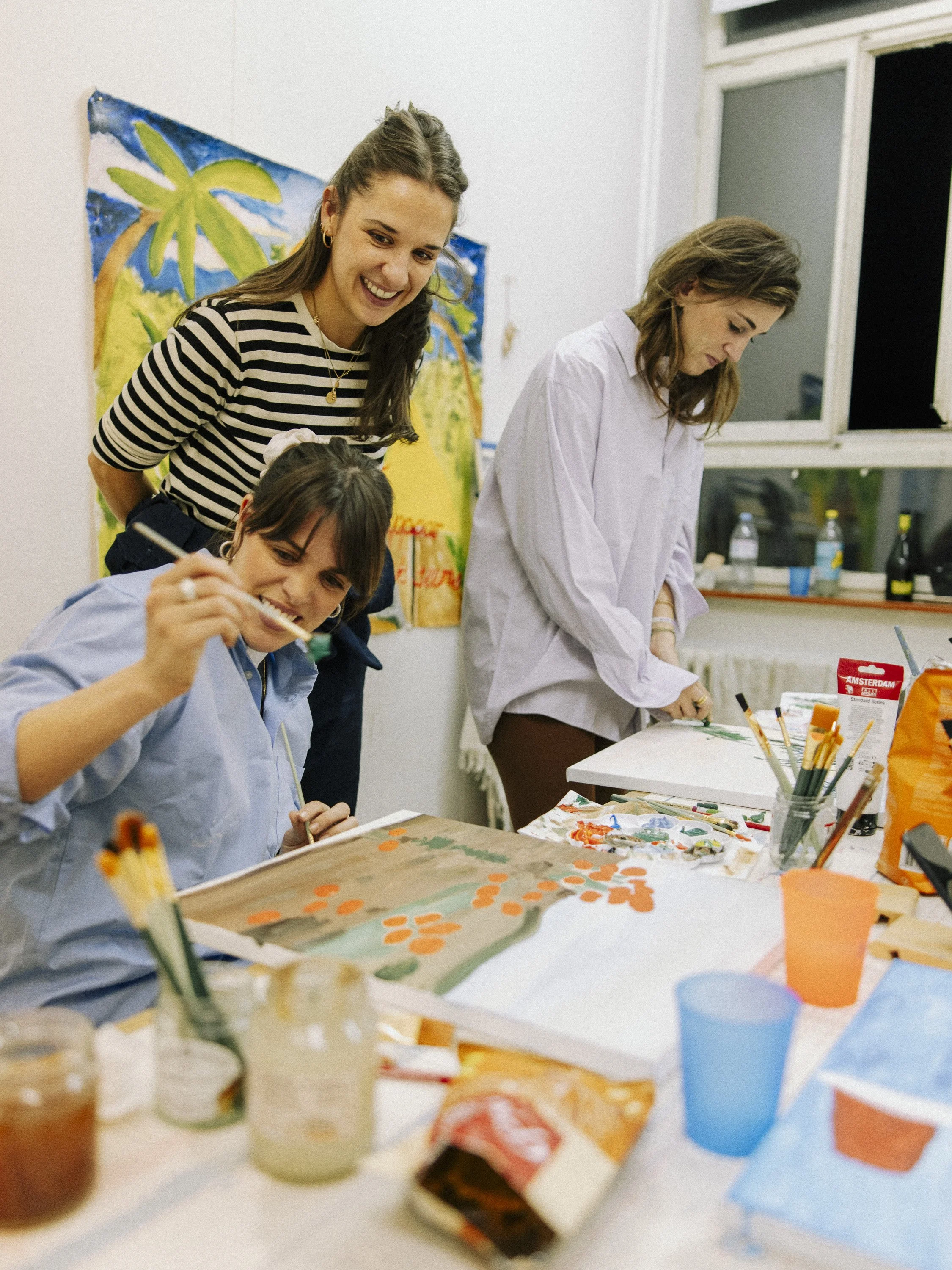 Three women at a table painting and creating artwork, one woman standing behind two women who are sitting and painting. The table is covered with paints, brushes, and art supplies, and there is a colorful painting of a tree on the wall in the background.