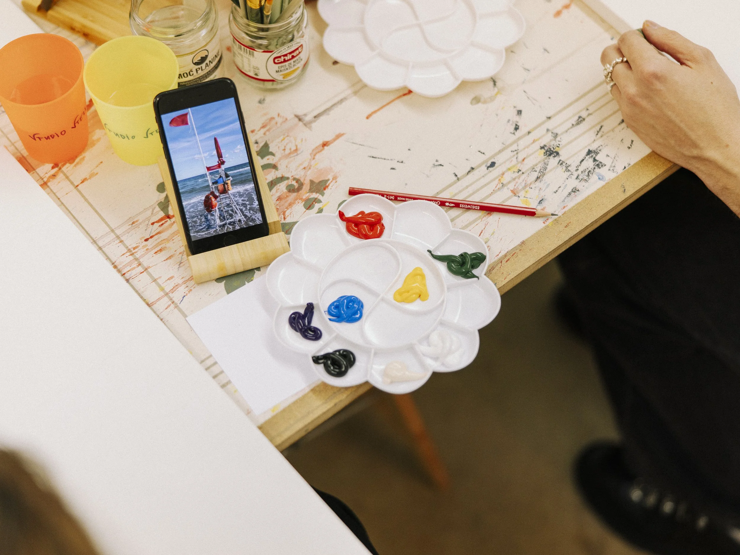 A tabletop with art supplies including a paint palette with blobs of red, blue, yellow, green, black, purple, and white paint, a paintbrush, and a smartphone displaying a photo of a boat on the water. There are also two colorful cups, jars, and a floral paper under the supplies.