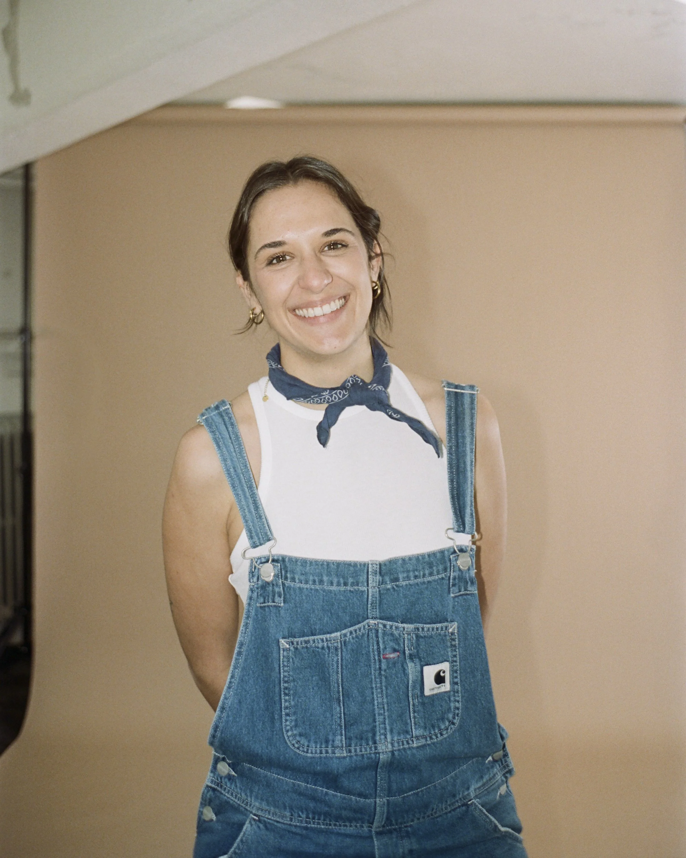 A smiling woman wearing denim overalls, a white tank top, a blue bandana around her neck, and gold earrings stands in front of a beige background.