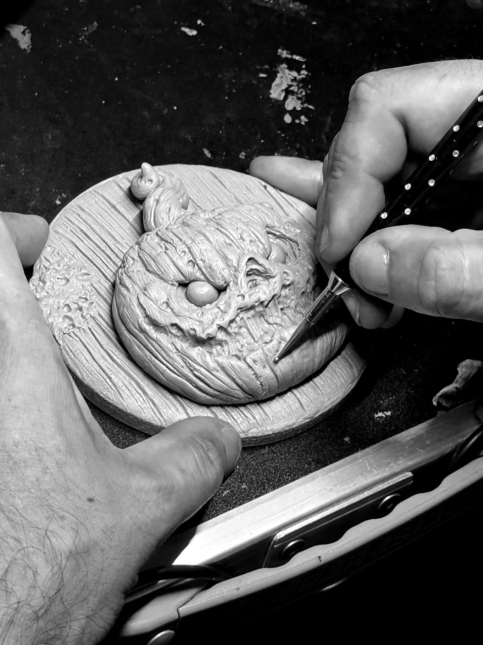 Une main tient un outil de sculpture et travaille sur une sculpture en argile d'un visage humain. La scène est en noir et blanc.