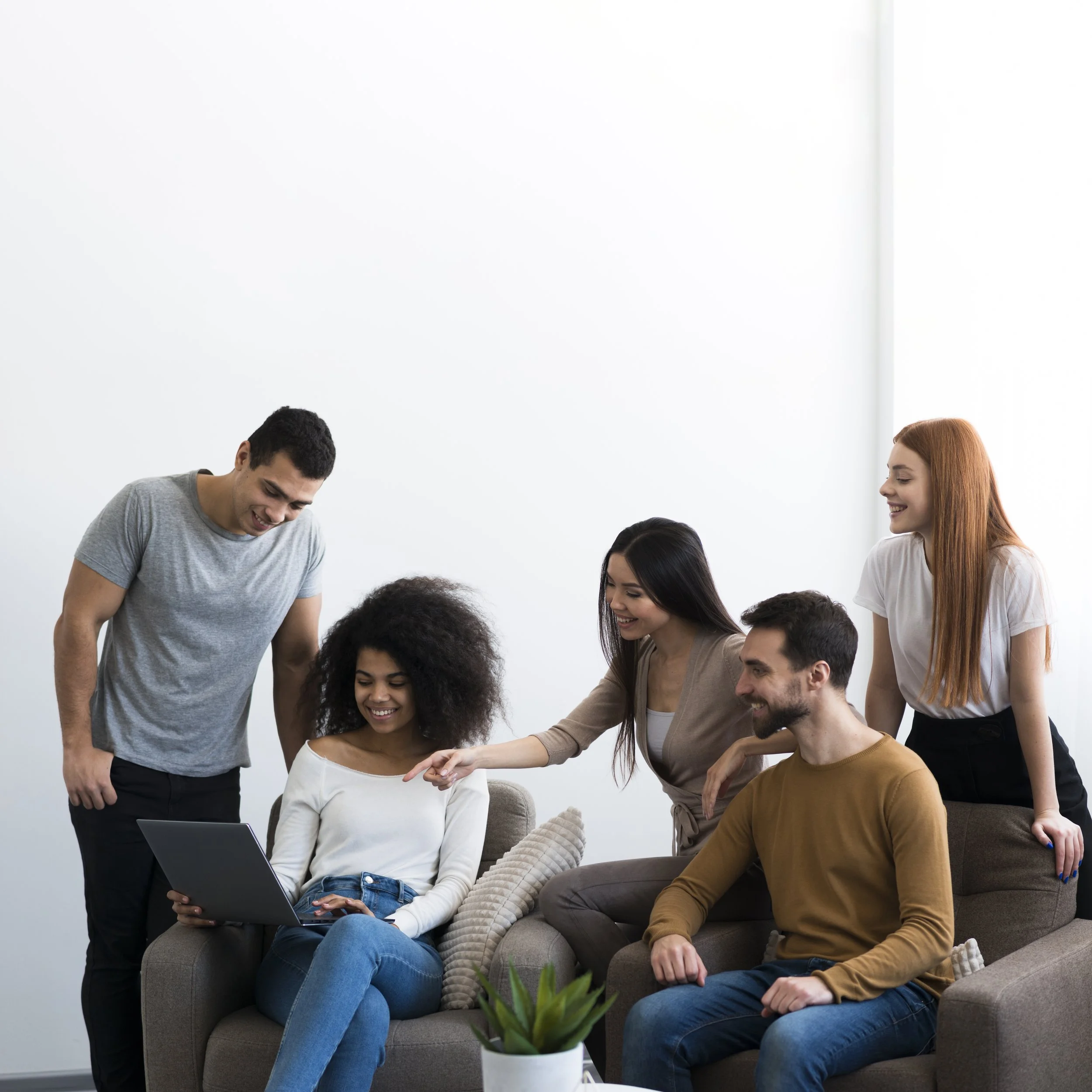 Group of five young adults gathered around a laptop, smiling and engaging in a conversation in a bright, minimalistic living room.