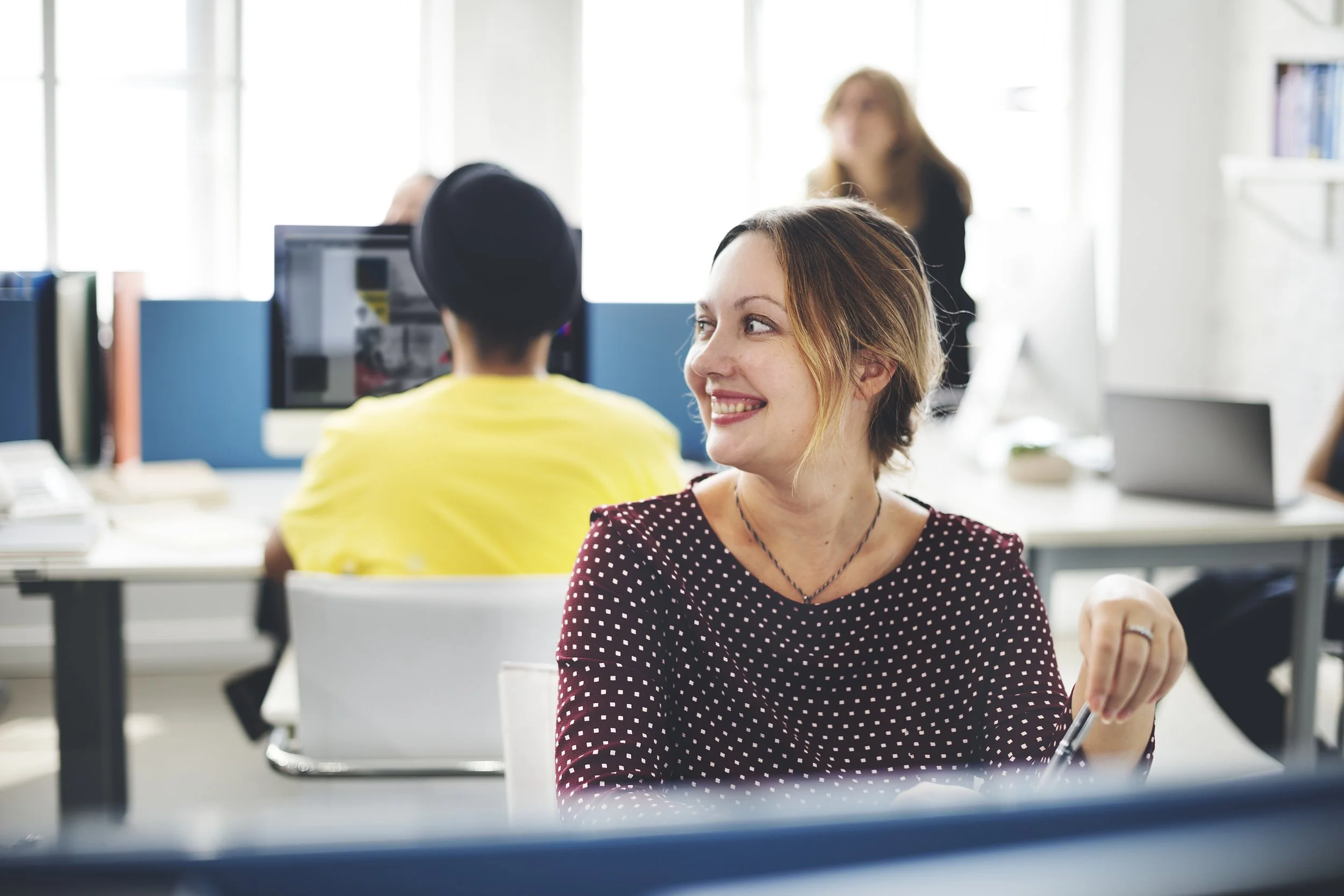 A woman smiling and looking to her right in an office, with coworkers working at desks in the background.