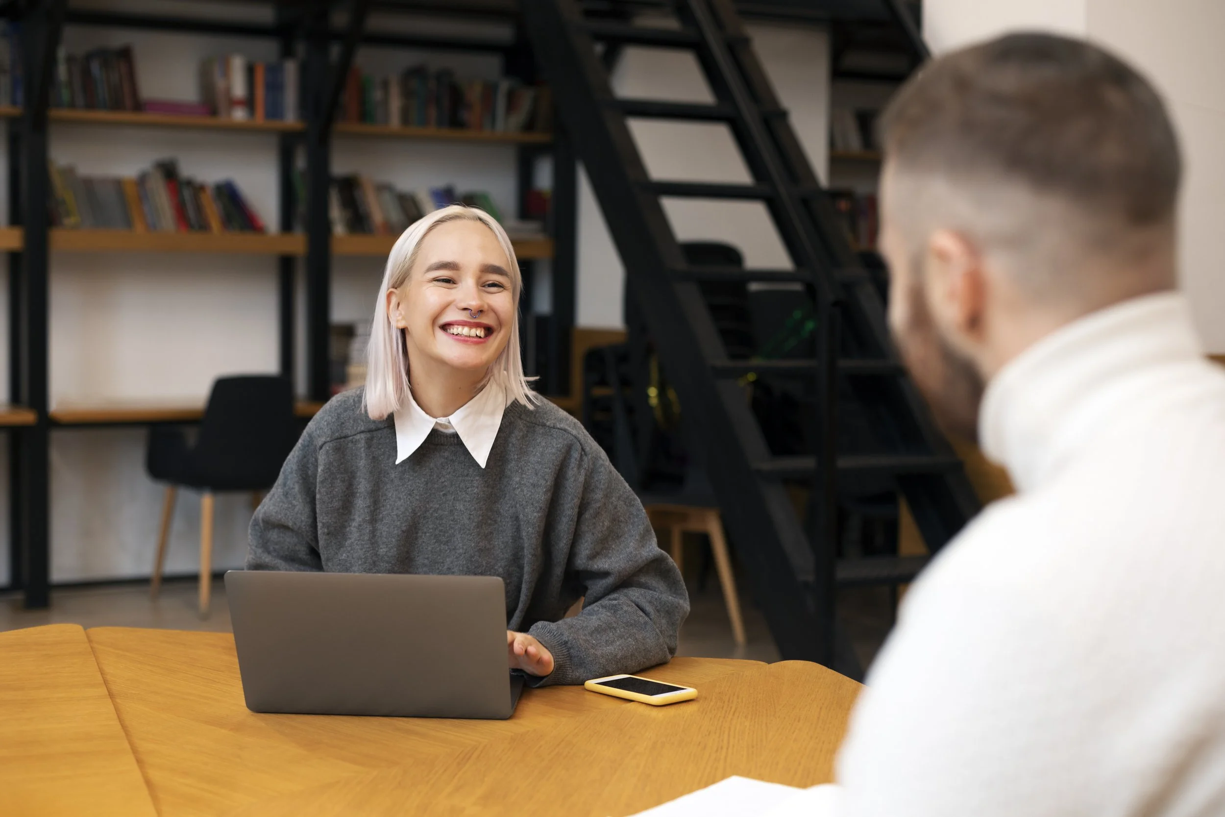 A woman with blonde hair and a gray sweater smiling during a conversation in a modern, cozy room or office with a bookshelf and black staircase in the background.
