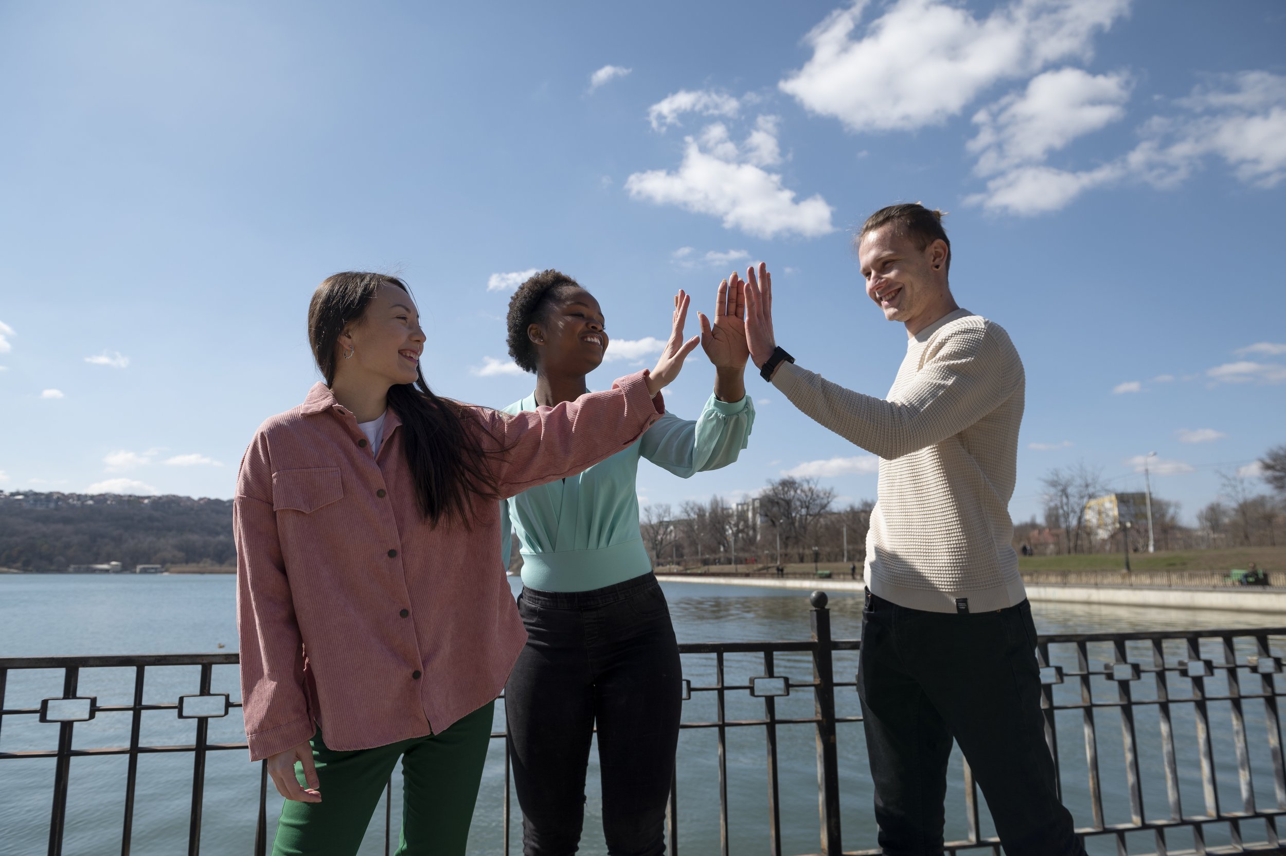 Three people giving each other high fives outdoors near a body of water on a sunny day.