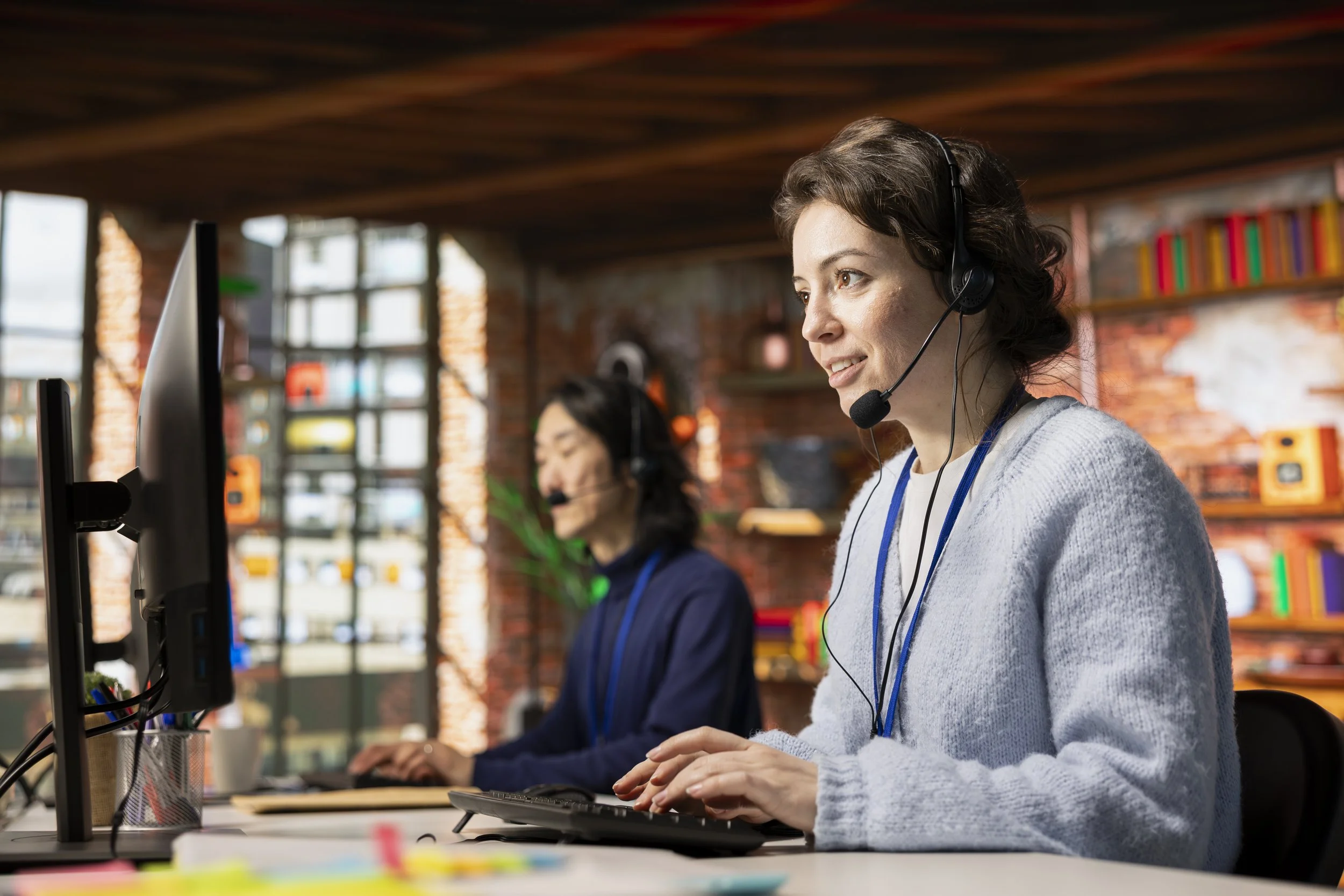 Two women wearing headsets working at computers in an office with a brick wall and large windows in the background