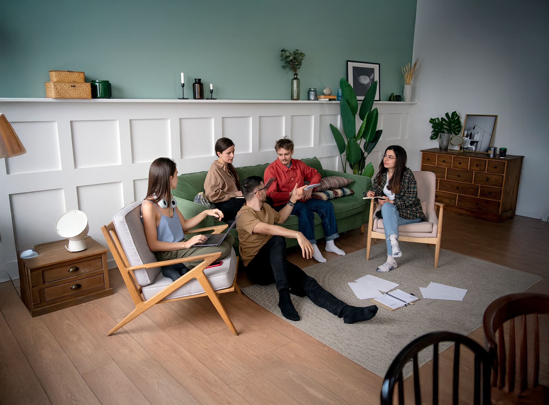 Five young adults with notebooks, tablets, and a laptop in a cozy living room with green walls, a white paneled wall, wooden furniture, and plants, engaged in discussion.