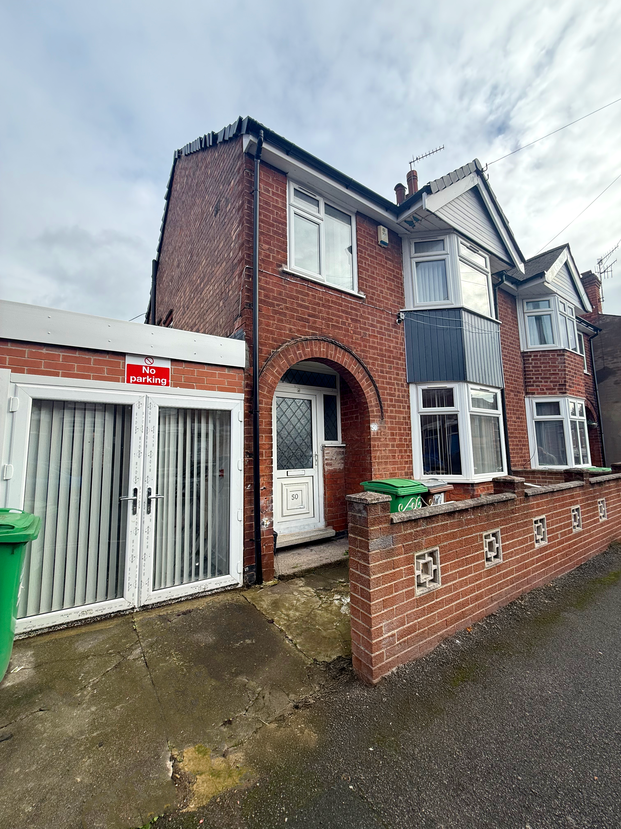 A brick house with a white front door, bay windows, and a small front yard enclosed by a brick wall. There is a 'No Parking' sign on a white gate and green trash bins outside.