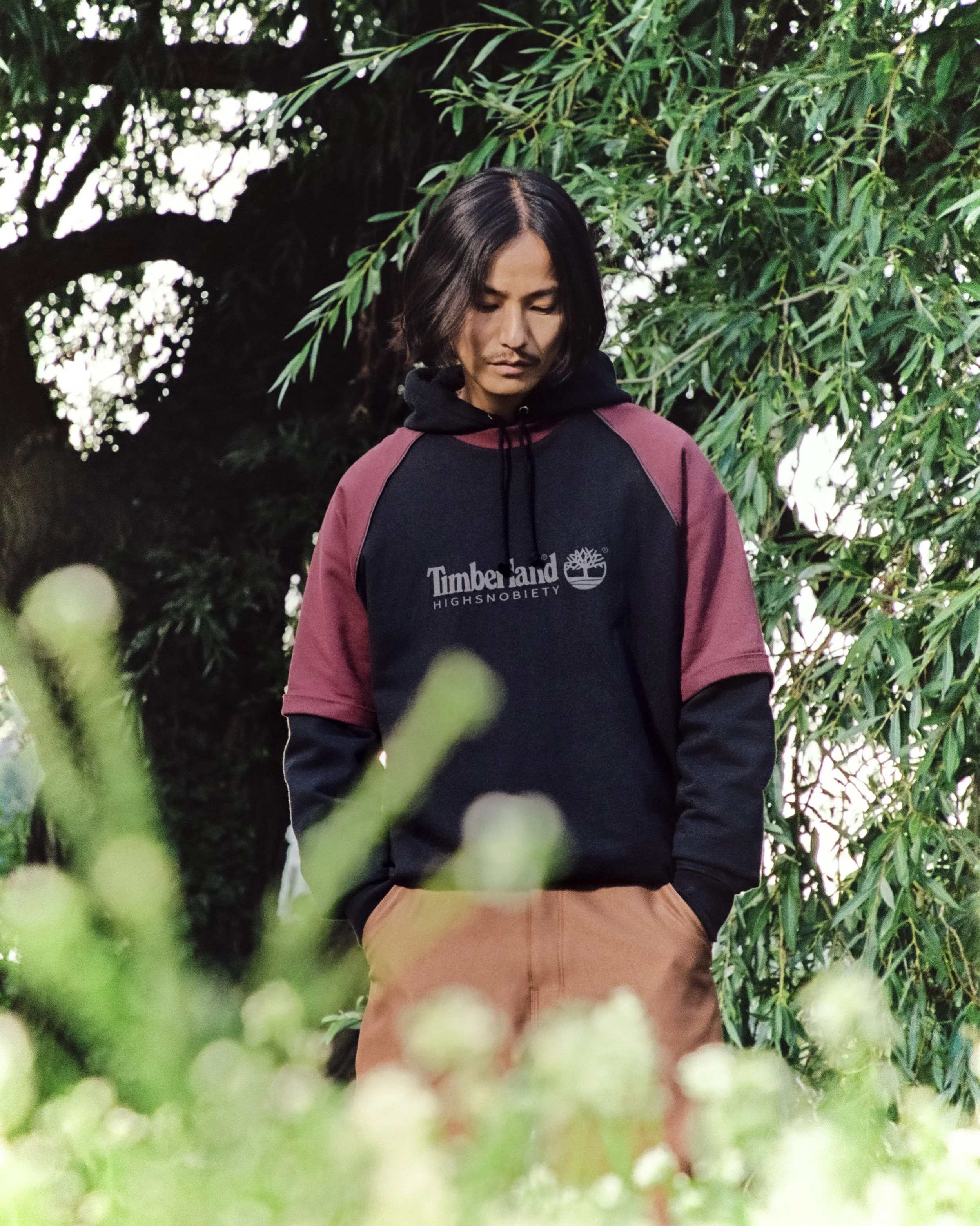 A young man with short black hair wearing a black, maroon, and gray Timberland hoodie and brown pants stands outdoors amongst green foliage, looking down with a thoughtful expression.