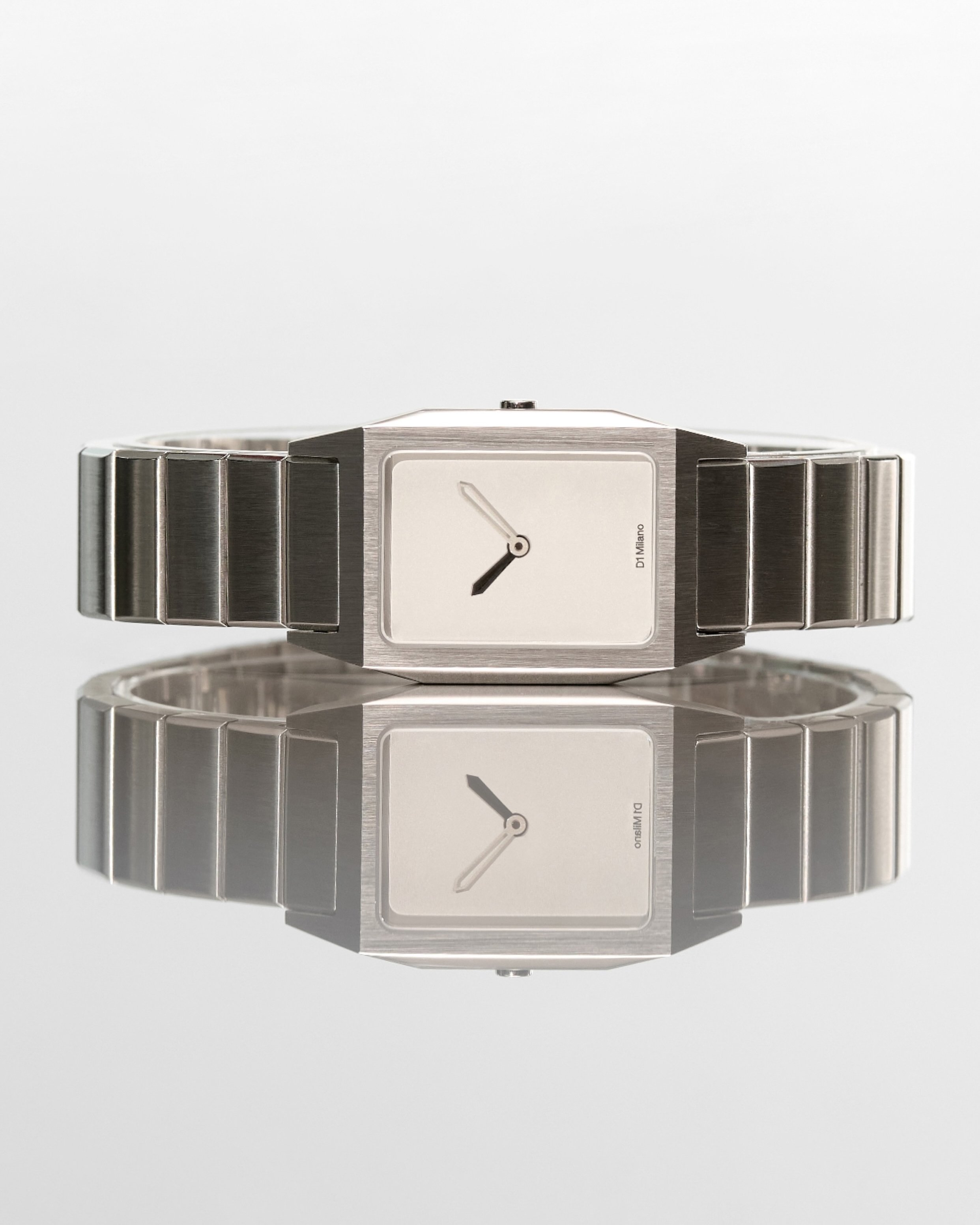 A silver wristwatch with a rectangular face and metal band, reflected on a mirror surface.