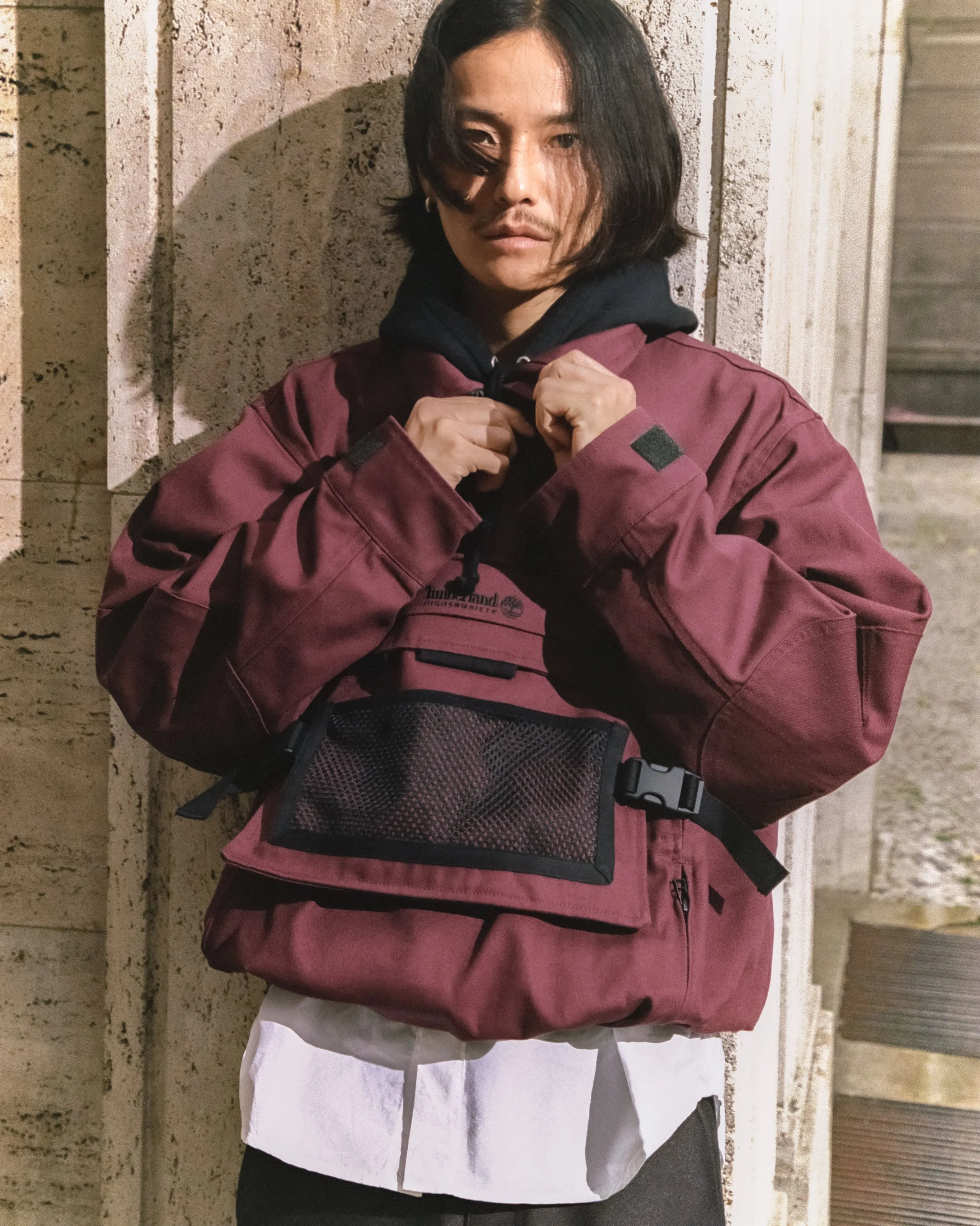 A person with long dark hair, wearing a maroon jacket and a black hoodie, standing outdoors at night against a concrete wall, adjusting their zippered hoodie.