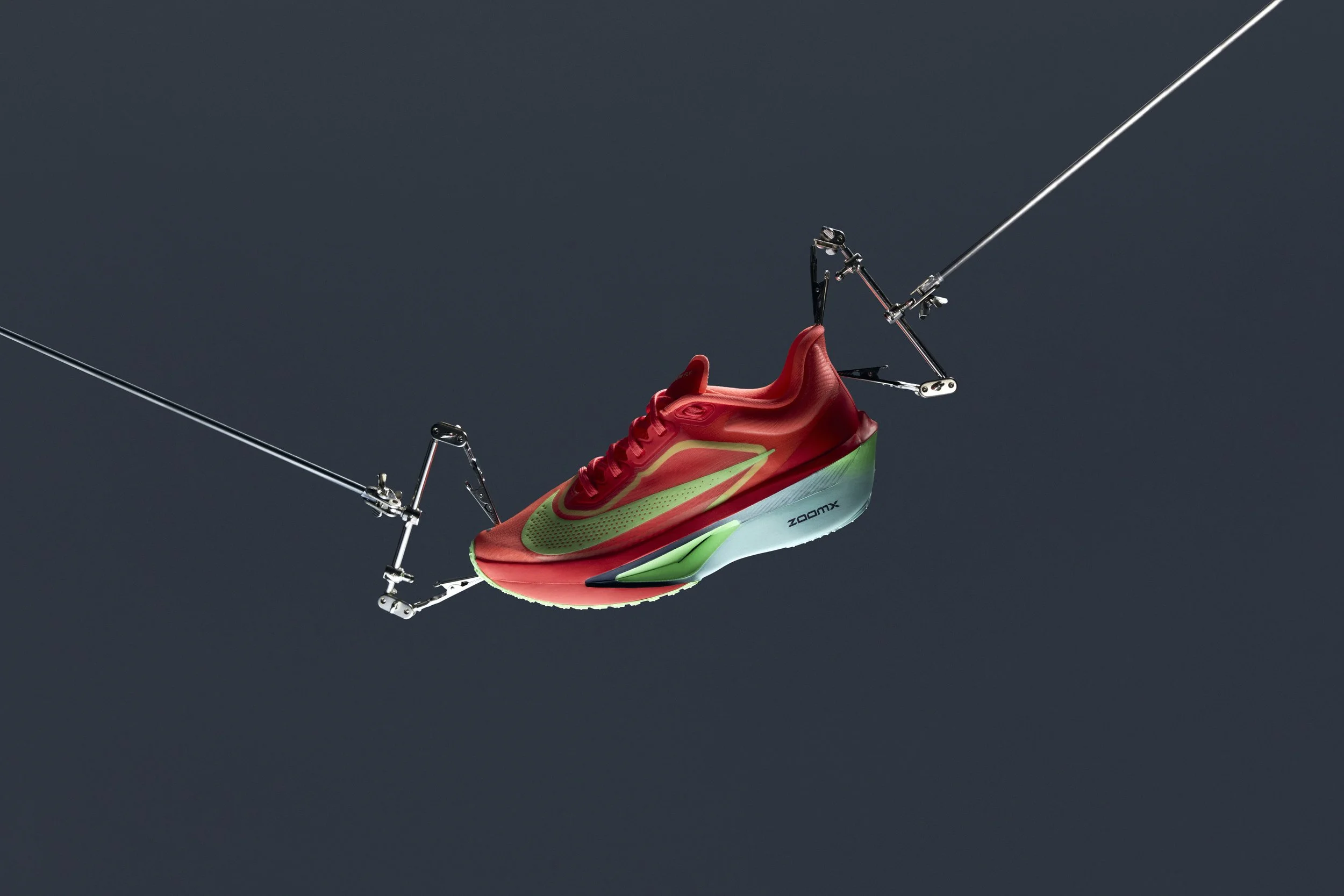 A red and green Nike running shoe suspended by cables against a dark background.