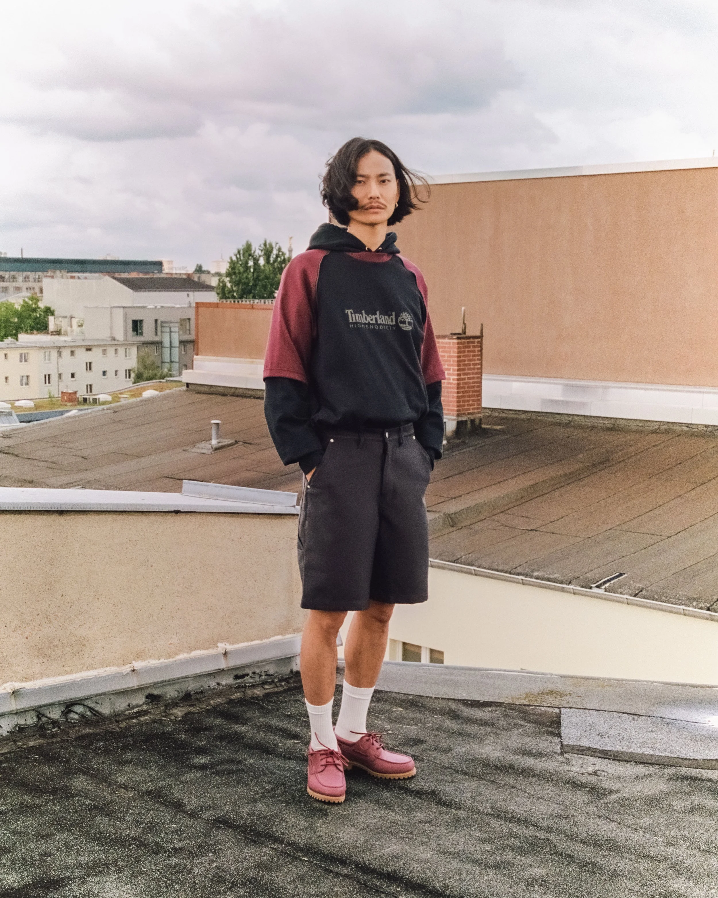 A young man standing on a rooftop in an urban area wearing a Timberland hoodie, dark shorts, white socks, and pink shoes, with cloudy skies overhead.