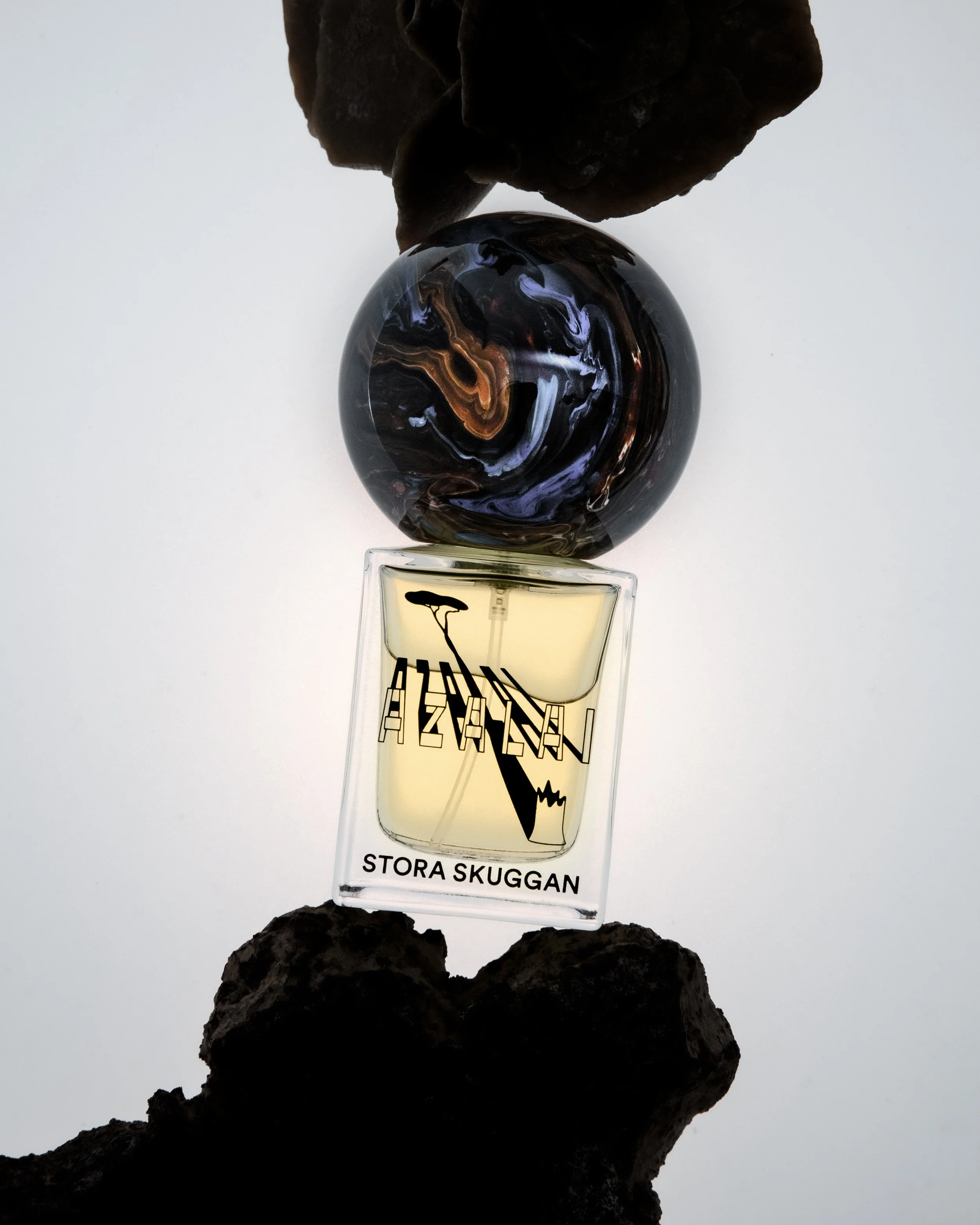 Flat lay of a perfume bottle labeled 'Stora Skuggans' with a black marble sphere and dark rocks arranged on a light background.