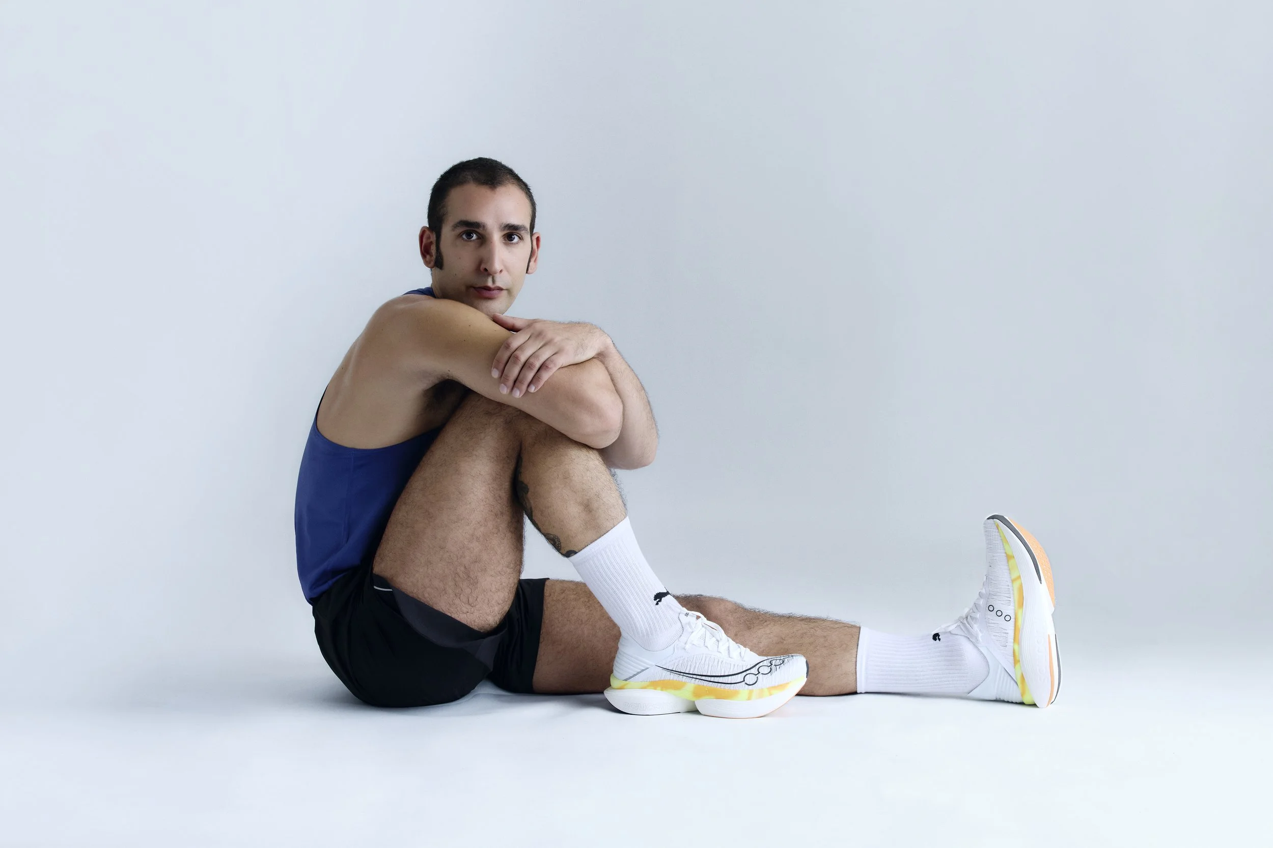Man sitting on the floor with arms around his knees, wearing a blue tank top, black shorts, white socks, and white athletic shoes, against a plain light background.